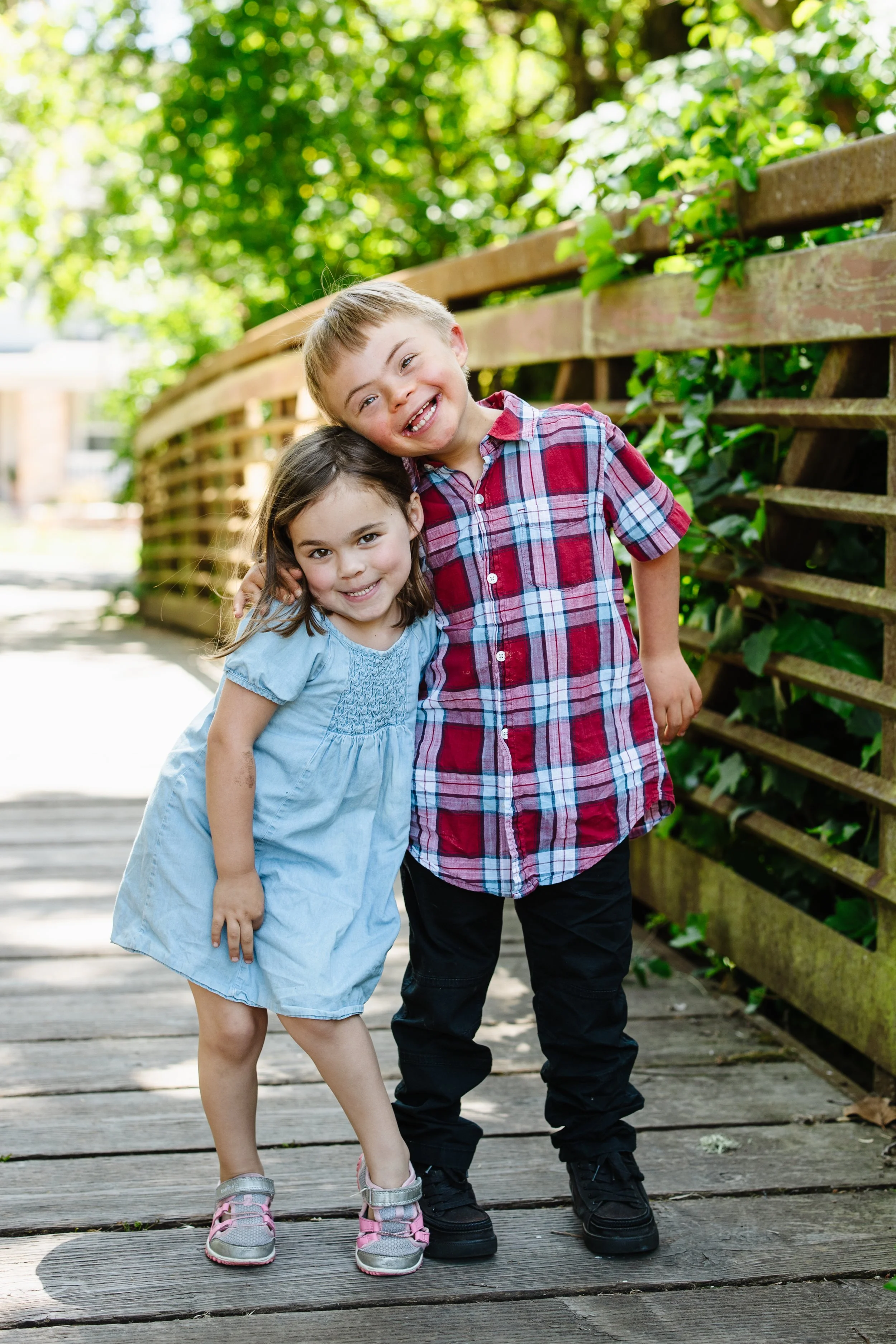Two children, a girl and a boy, happy and smiling, standing on a wooden bridge surrounded by greenery.
