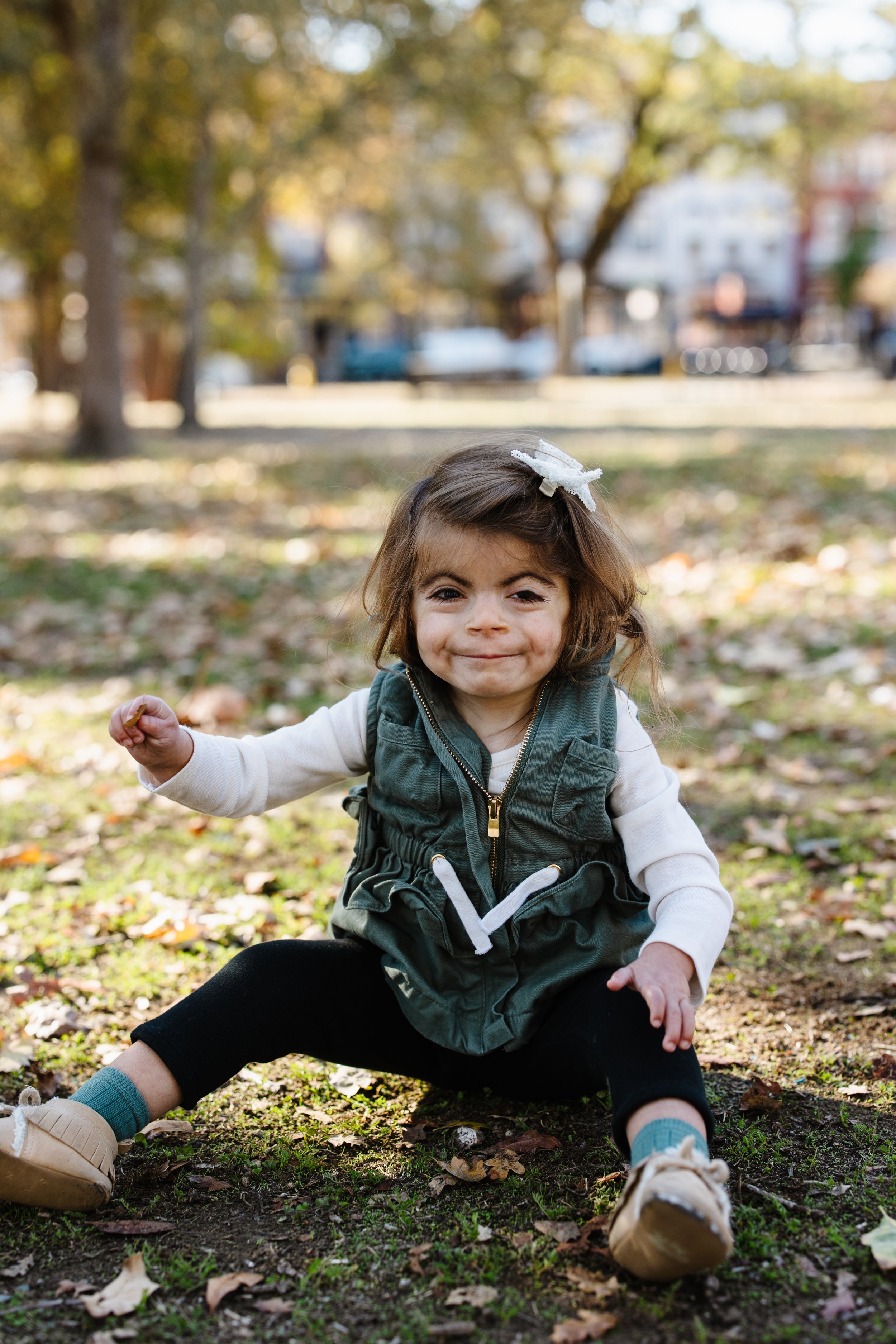 Young girl sitting on the ground in a park, smiling and making a fist. She has brown hair, a white bow, a green vest, beige shoes, and black pants, with autumn leaves on the ground.