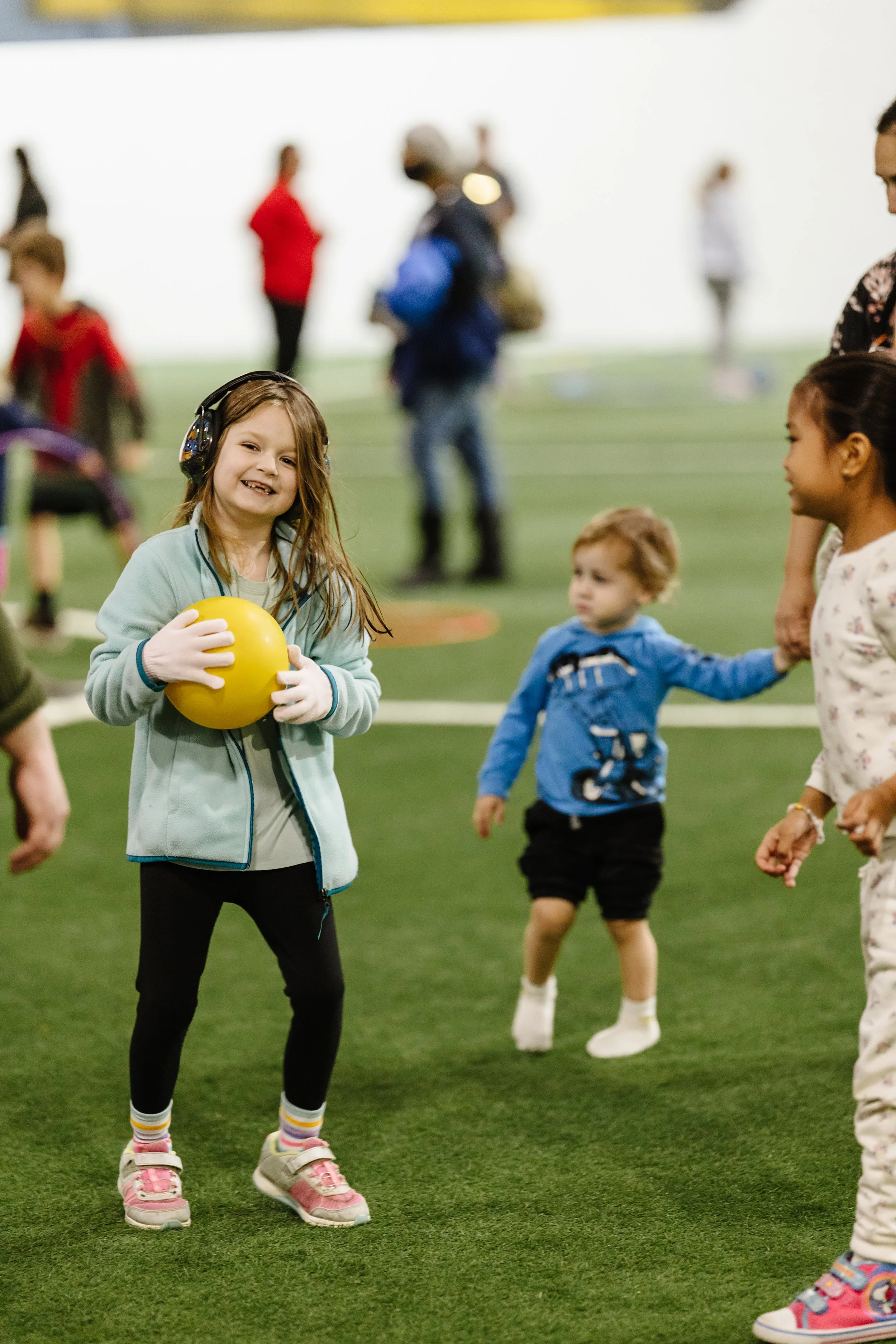 Group of children playing on an indoor field, with a girl in the foreground holding a yellow ball, smiling, wearing headphones and colorful sneakers.