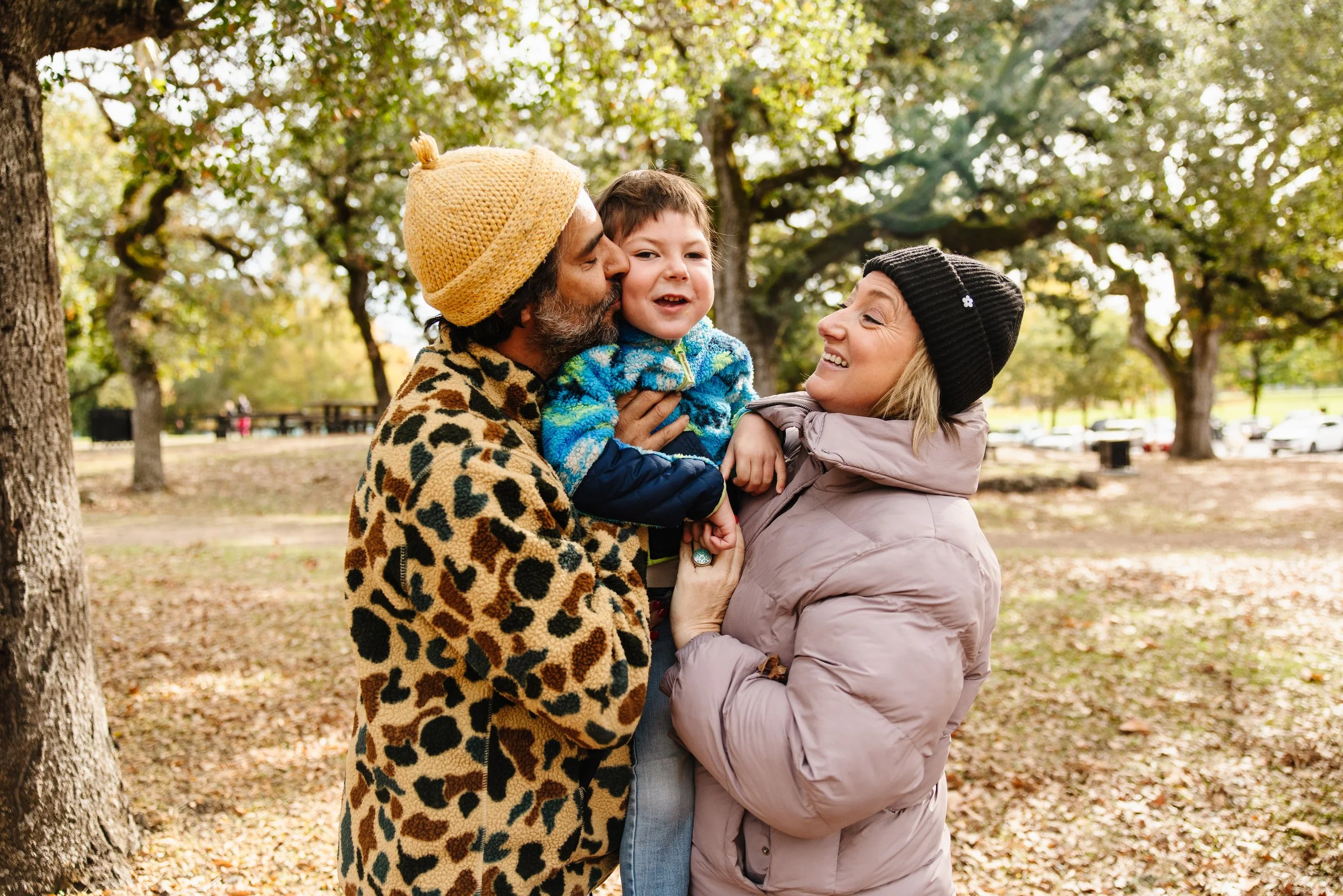 A family of three, including a man, a woman, and a young boy, enjoying a moment together in a park during autumn. The man is wearing a yellow knit beanie and a leopard print coat, the woman is wearing a black beanie and a light pink puffer jacket, and the boy is being held by both, wearing a blue patterned jacket. The family is smiling and sharing a tender moment, surrounded by trees with green and yellow leaves and fallen autumn leaves on the ground.