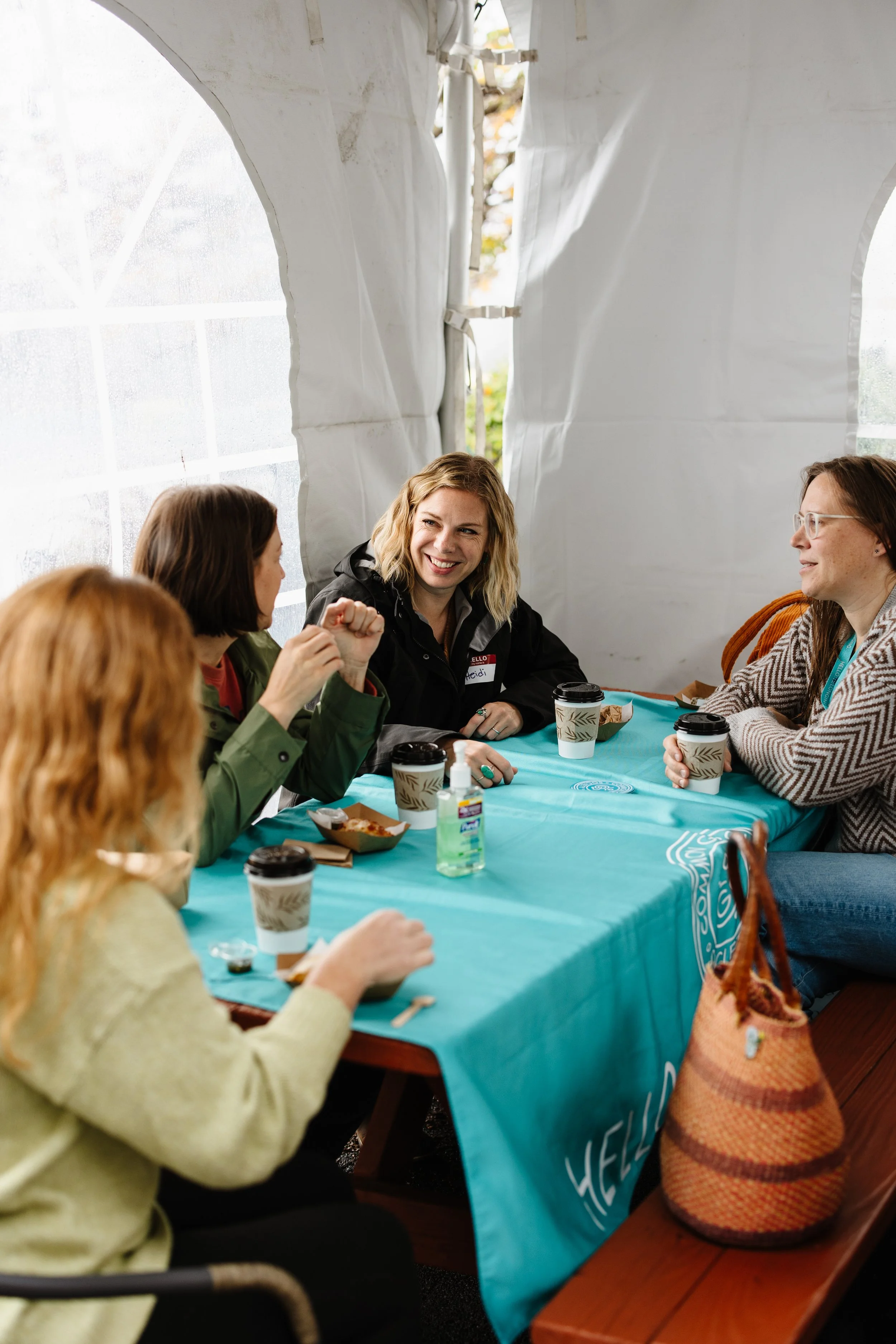 Five women are sitting at a table inside a tent, having a conversation and smiling. They each have cups of coffee or tea and some snacks. The table is covered with a blue tablecloth, and a hand sanitizer is on the table. One woman is wearing a green sweater, and another has a striped sweater. There is a woven bag on the bench next to one of the women.