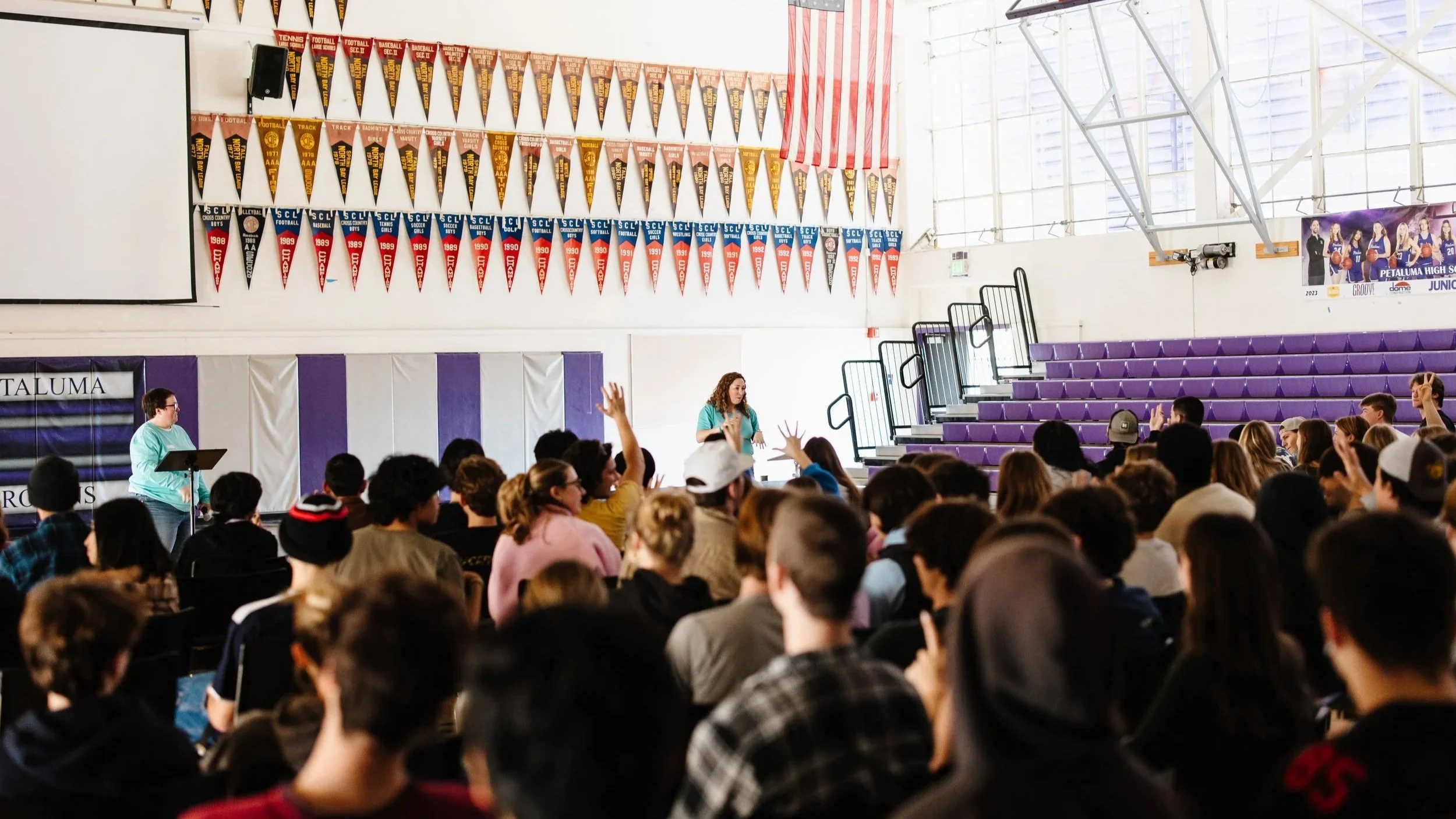A school assembly in a gymnasium with students sitting on chairs facing a speaker at a podium. Banners hang on the wall, and purple bleachers are on the right side. The speaker is a woman with curly hair, wearing a teal top, addressing the crowd. Some students have their hands raised.