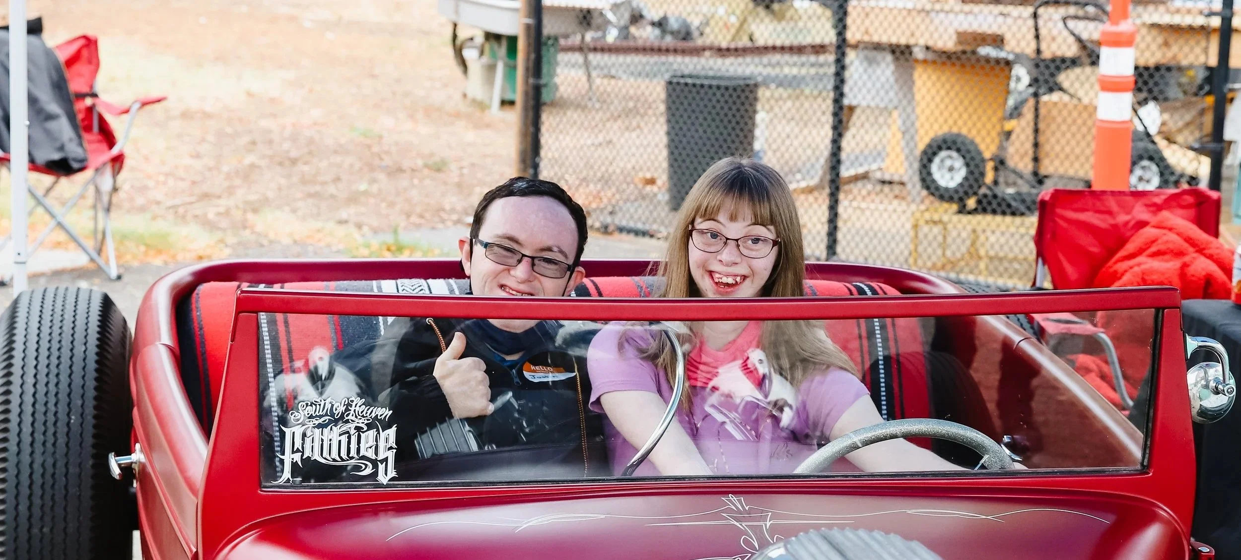 A boy and a girl sitting in a red vintage-style car with a glass windshield. The boy, wearing glasses and a black jacket, is giving a thumbs-up and winking. The girl, with long light brown hair and glasses, is smiling widely. There are trees and outdoor equipment in the background.