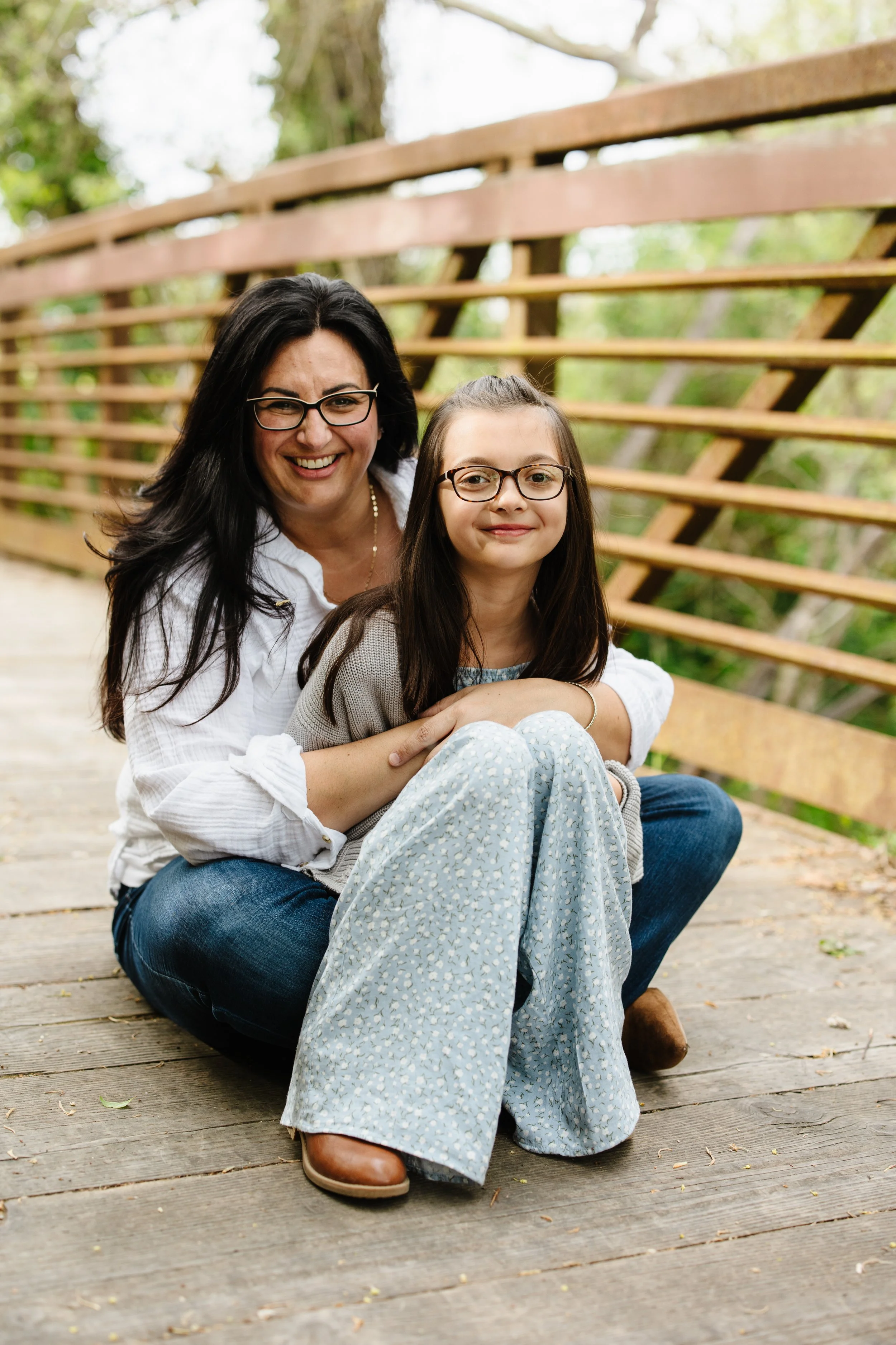 A woman and a young girl sitting on a wooden bridge outdoors, smiling at the camera, surrounded by green trees.