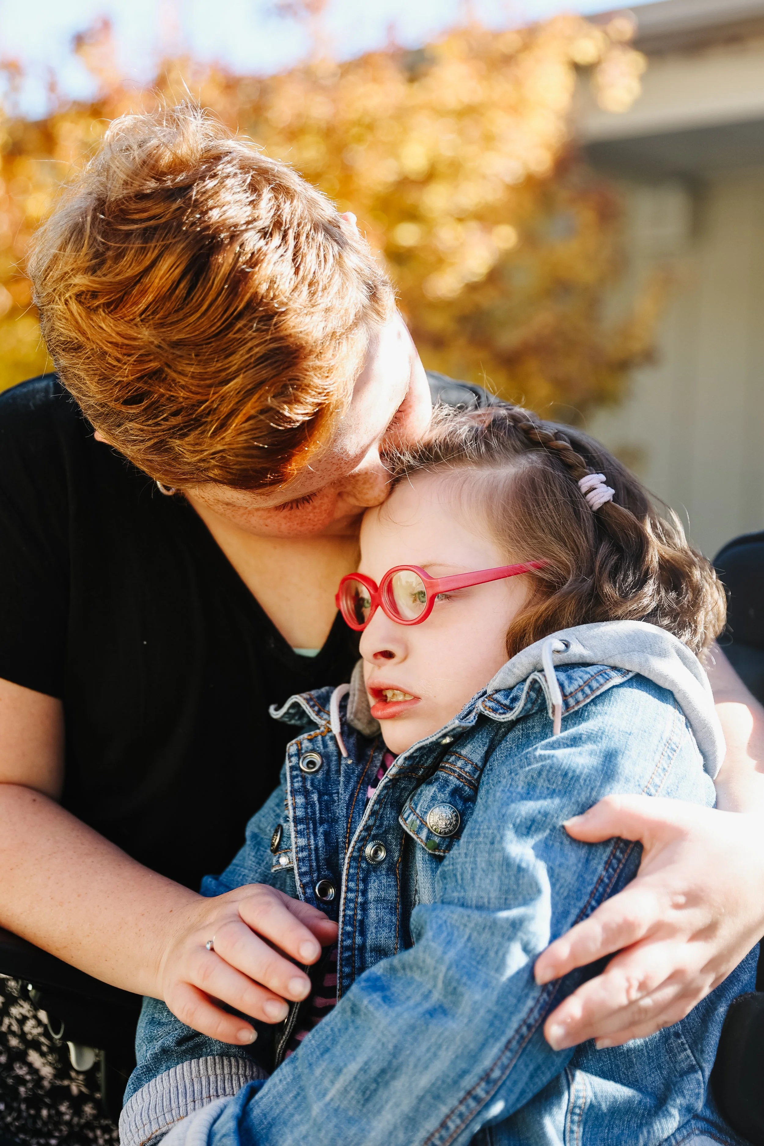 A woman with short reddish hair embraces a young girl with glasses and braided hair, outdoors on a sunny day with autumn-colored trees in the background.