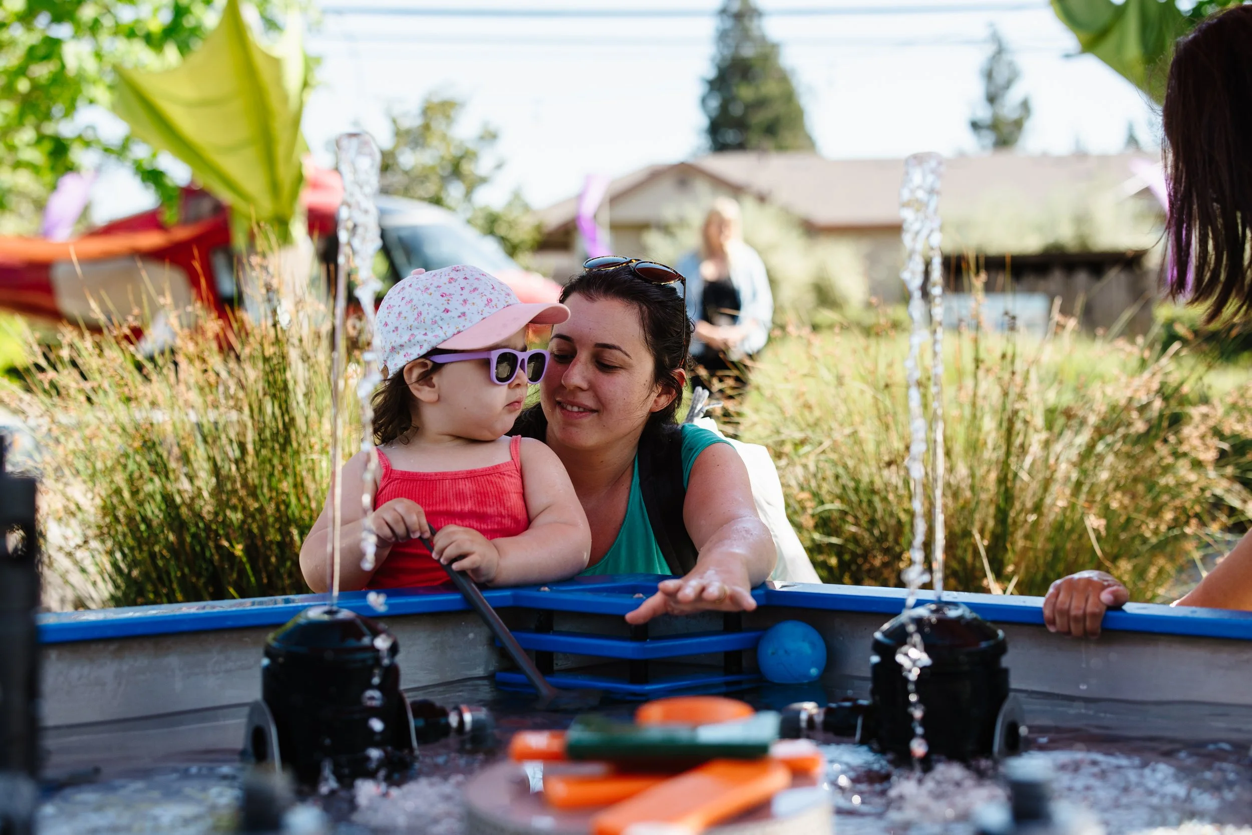 A woman and young girl playing with a small water table outdoors on a sunny day.