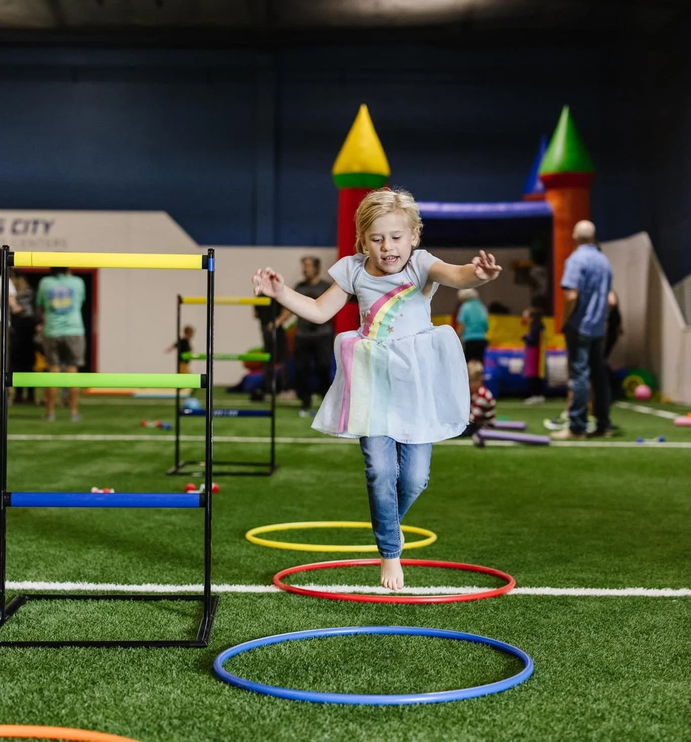 A young girl wearing a rainbow dress and blue jeans is jumping through colorful hula hoops in an indoor play area with artificial green turf. In the background, a bounce house shaped like a castle and other children and adults are visible.