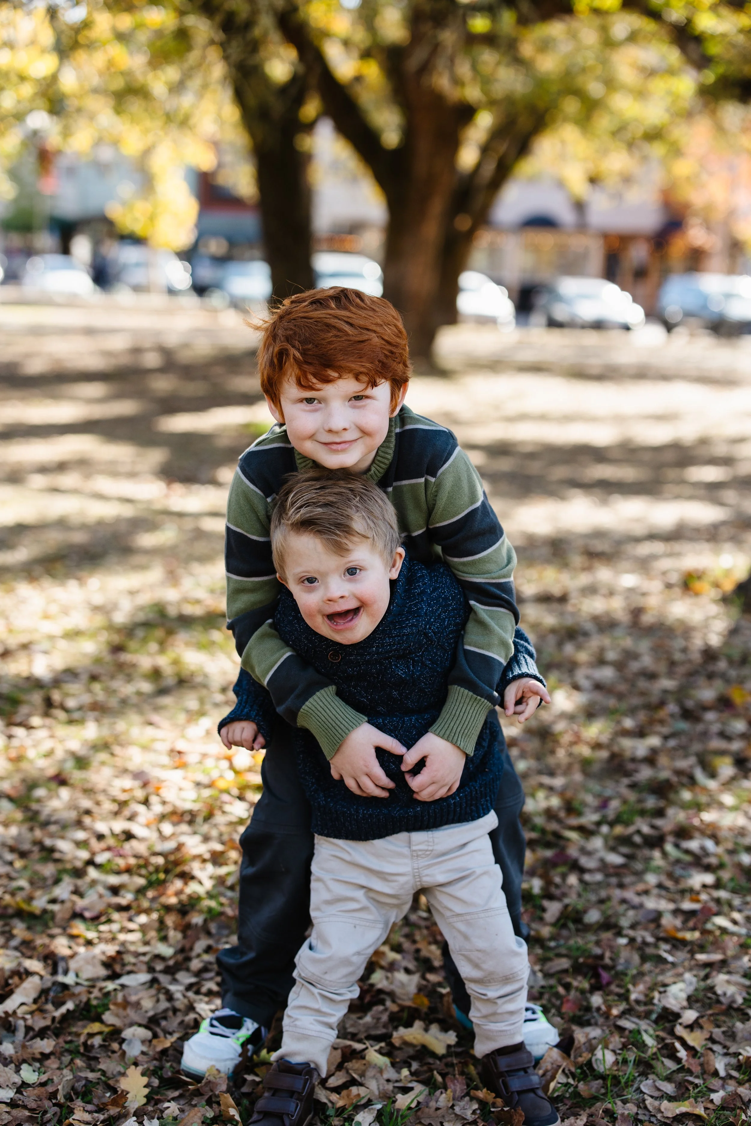 Two young boys playing outdoors in a park during autumn, with fallen leaves, trees, and parked cars in the background. The older boy is giving the younger boy a piggyback ride.