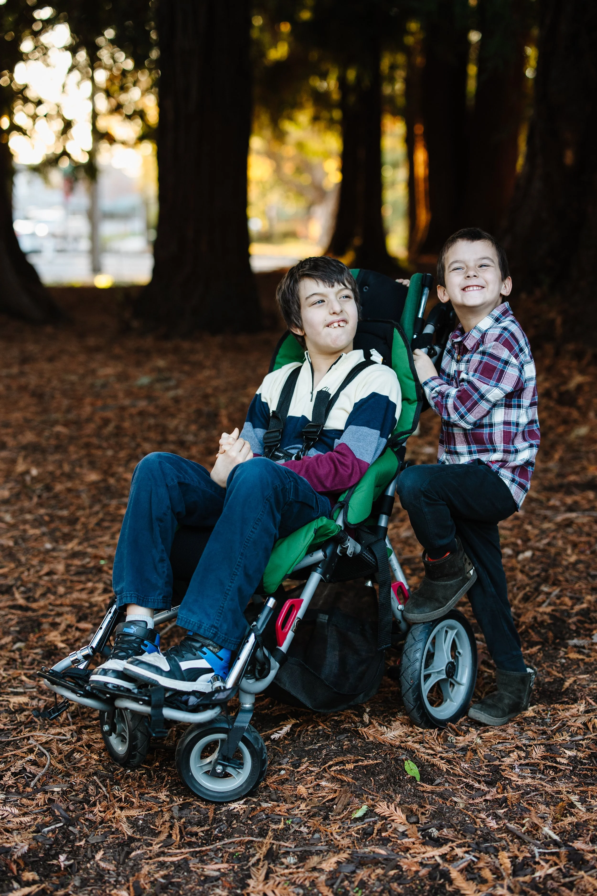 Two young boys, one sitting in a wheelchair and the other standing beside him, outdoors among trees with fall leaves on the ground, smiling and making silly faces.