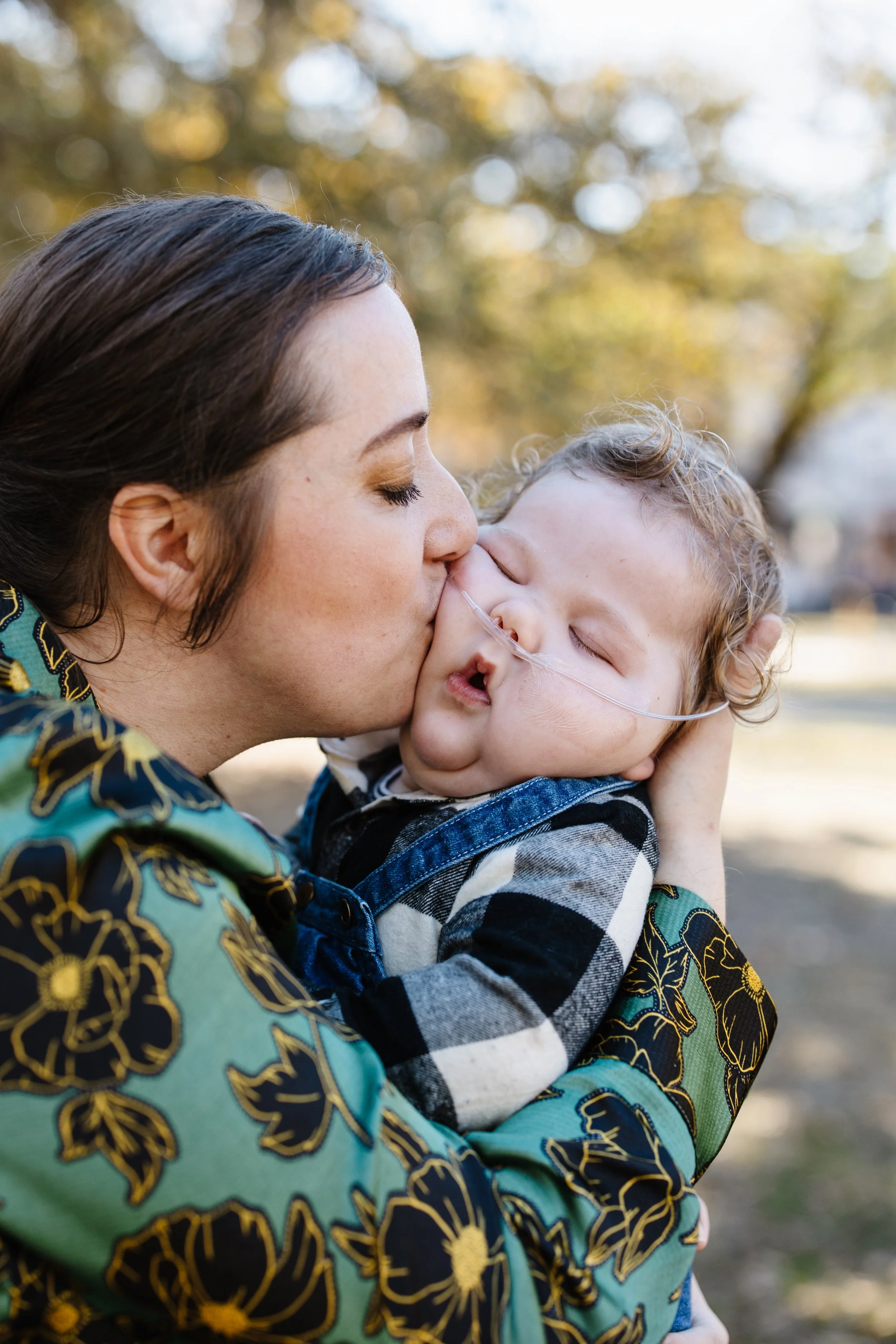 A woman with brown hair holding and kissing a young child with a nasal cannula outdoors during fall.