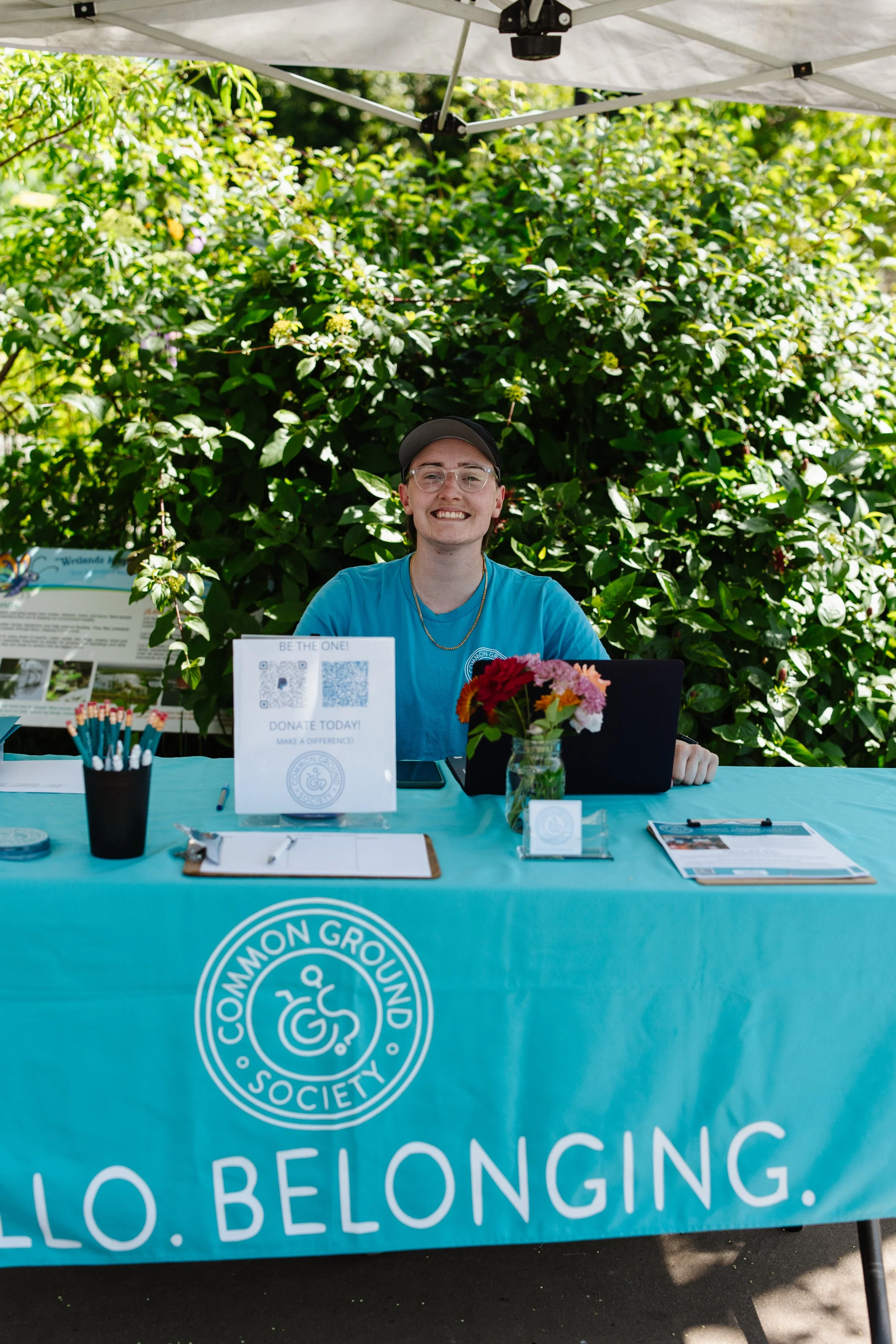 A smiling person sitting behind a table with a teal tablecloth at a community event, with a large green bush in the background. The table has a vase of flowers, informational signage, pens, and a laptop. The tablecloth has the logo and name of the 'Common Ground Society' and the words 'Hello. Belonging.'
