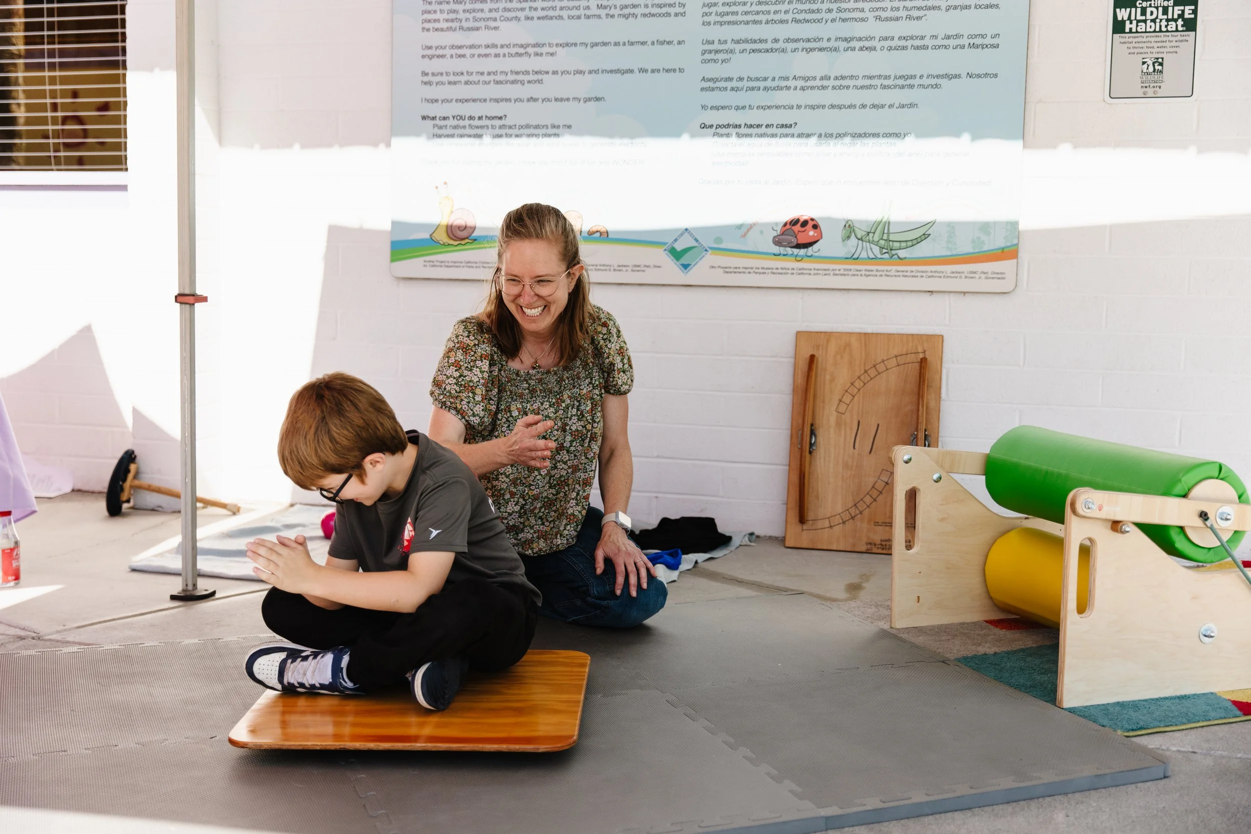 A young boy sitting on a wooden platform with his hands together in a prayer position, smiling, as a woman kneels beside him, laughing. They are in a room with educational posters and play equipment, including a green and yellow roller device, and a wooden balance board in the background.