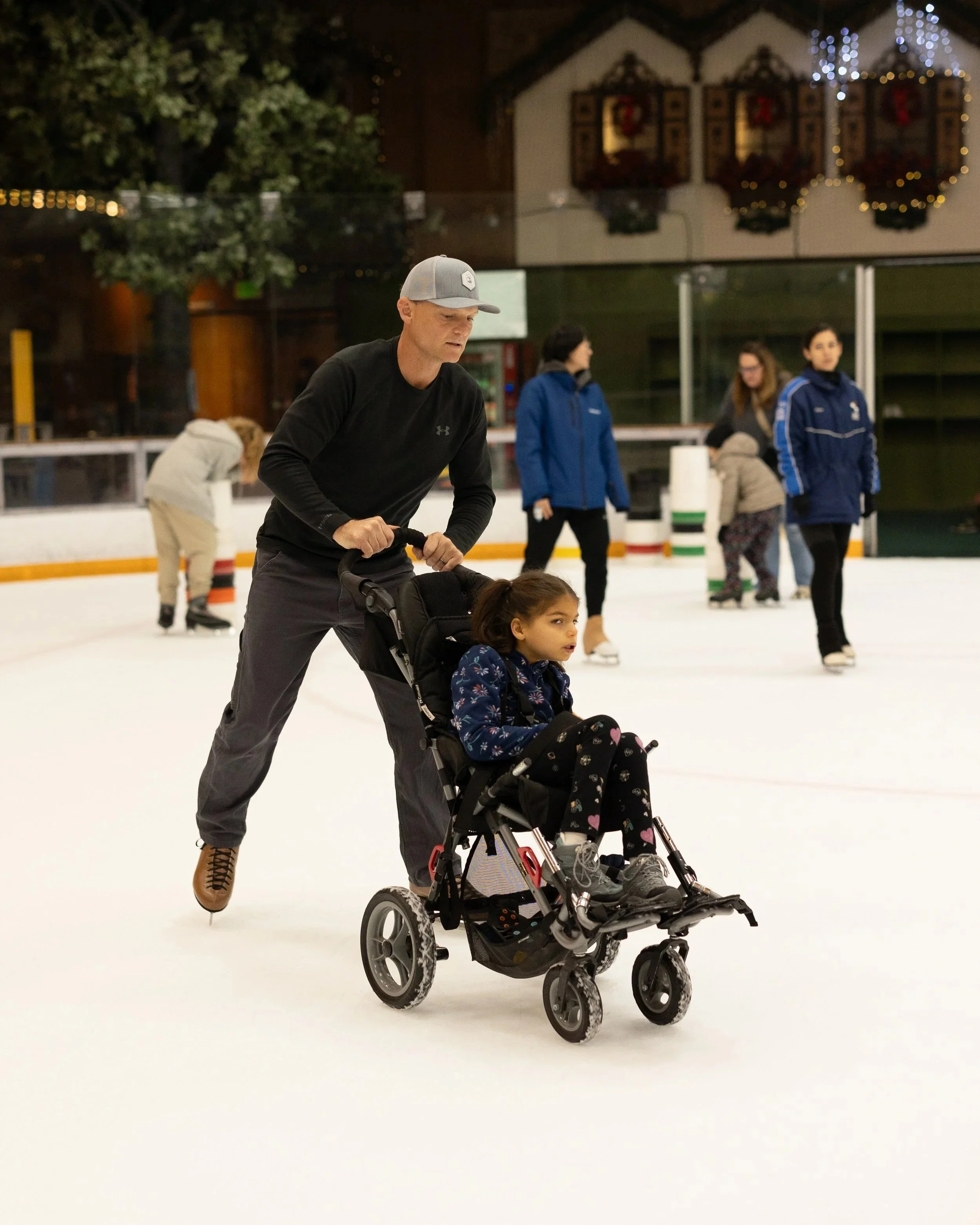 A man in a black shirt and gray cap pushing a young girl in a wheelchair on an ice skating rink. In the background, several people are skating and festive holiday decorations are visible in a decorated room.