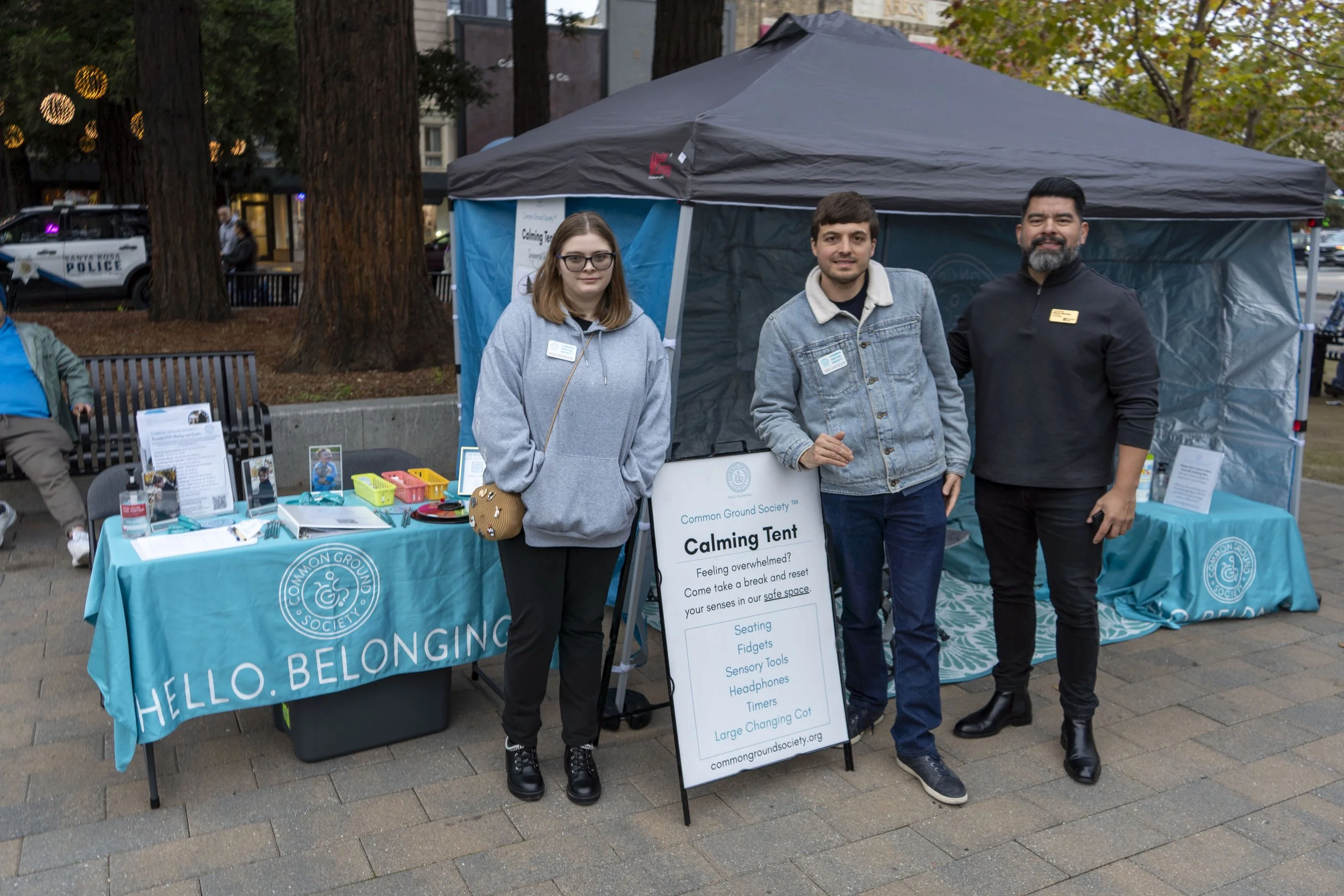 Three people standing in front of a booth at an outdoor event, with trees and city buildings in the background. The booth has a blue tablecloth with the name 'Common Ground Society' and a sign that reads 'Calming Tent' and lists activities like seating, fidgets, sensory tools, headphones, timers, and a large changing cot. The three people are smiling, two women and one man, all wearing casual attire.
