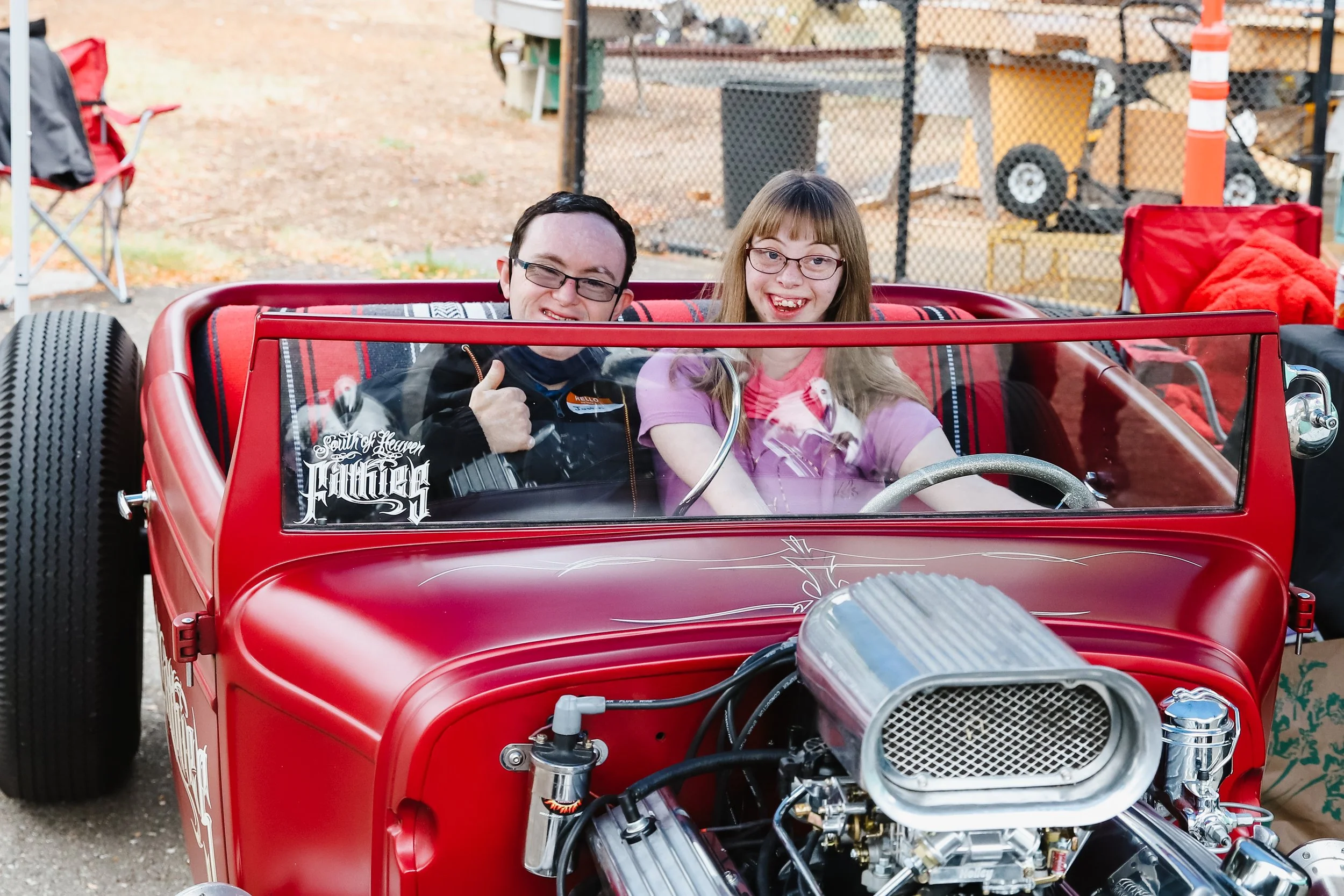 Two people in a red vintage car, smiling and giving a thumbs up, with a checkered interior and a large engine at the front, parked outdoors