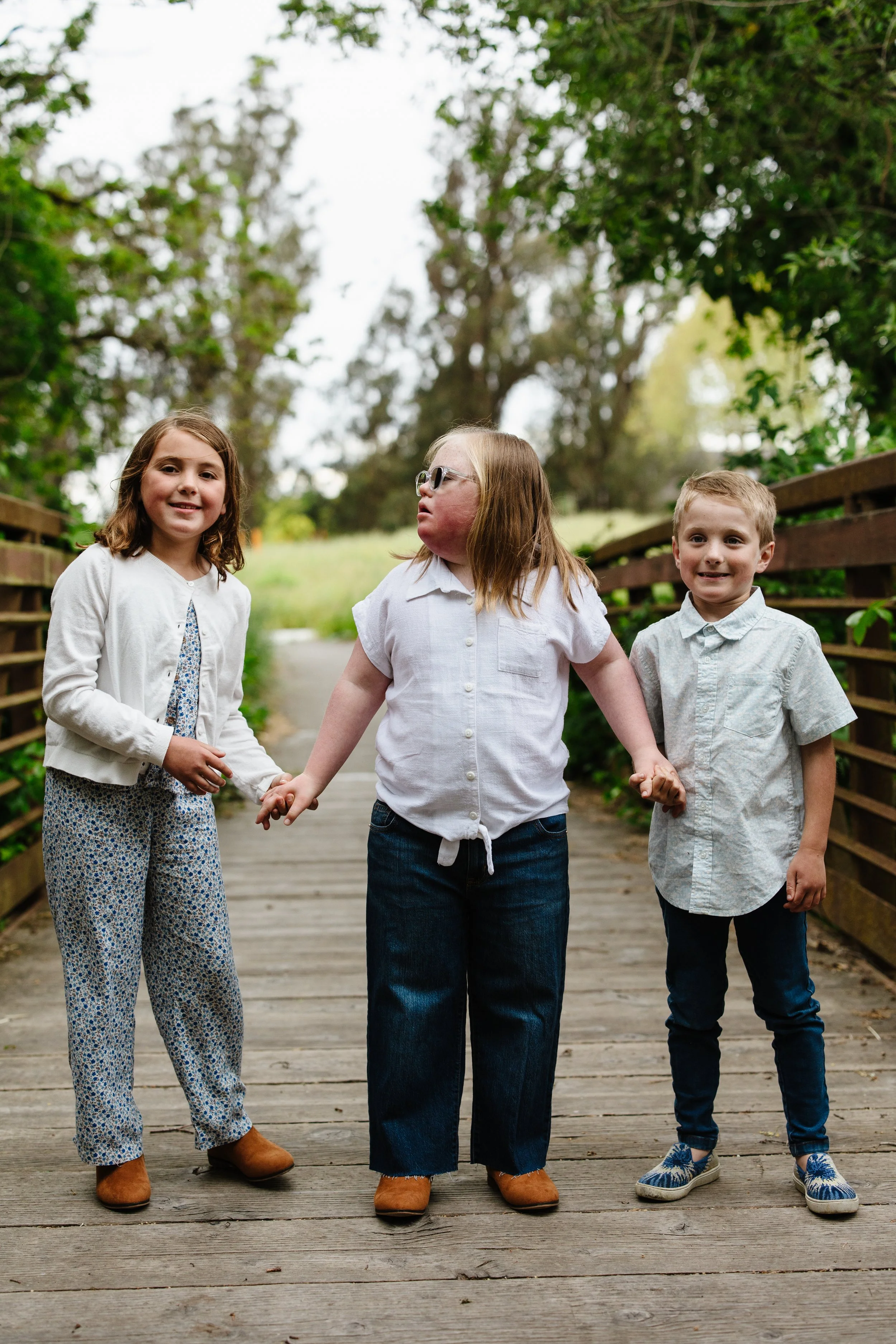 Three children holding hands on a wooden bridge outdoors surrounded by green trees.