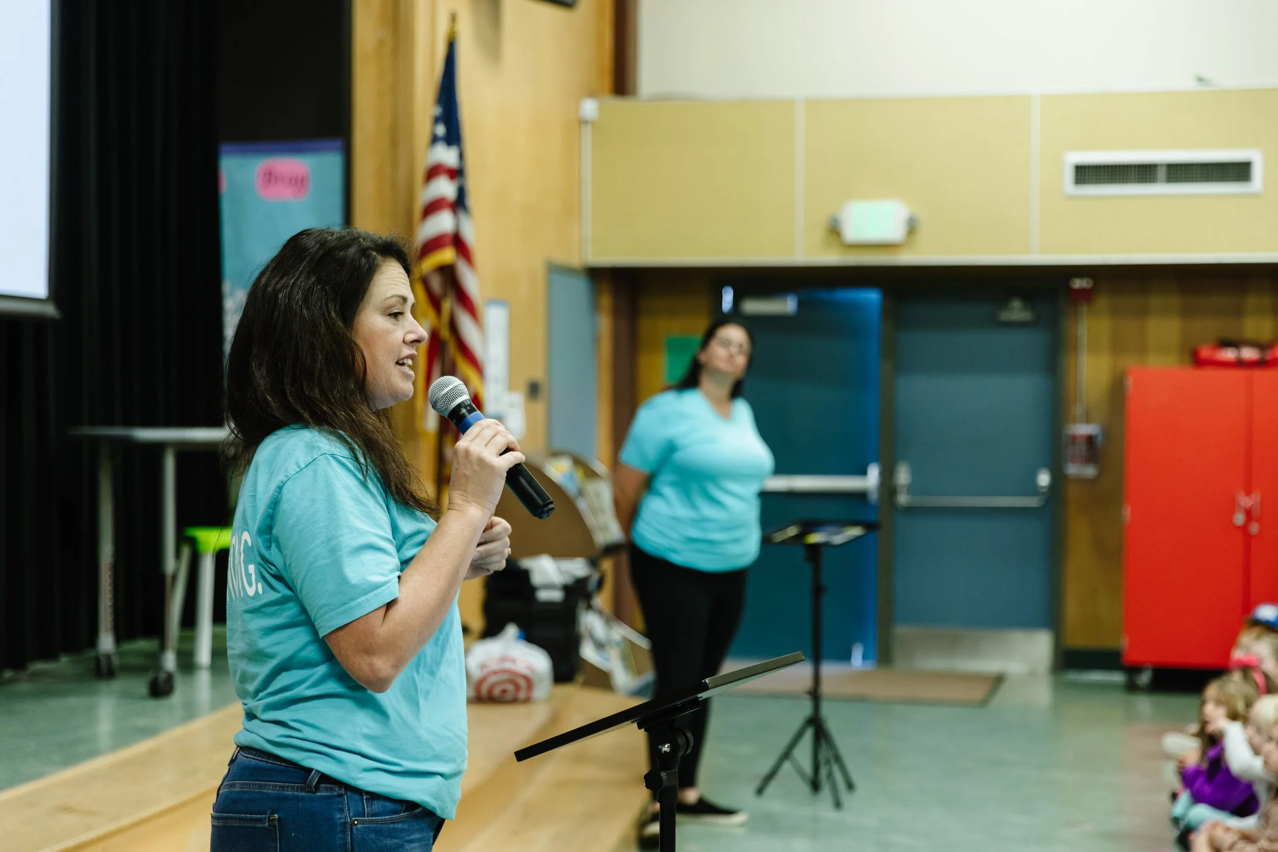 Common Ground presenter holding a microphone and speaking to children seated on the floor in an elementary school auditorium. A woman in a light blue shirt stands in the background near a door, with an American flag and classroom equipment visible.