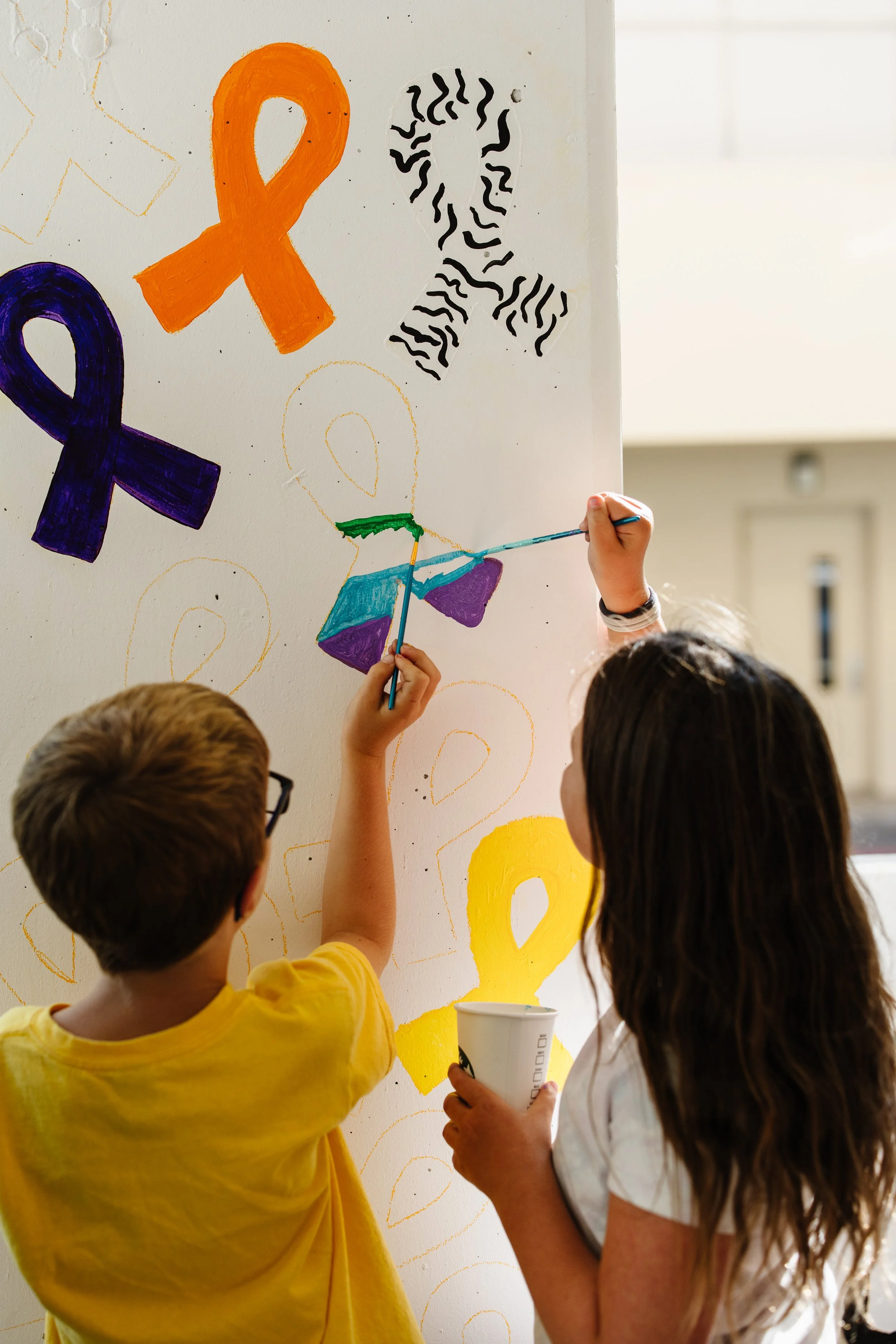 Two children, a boy and a girl, creating colorful artwork on a white wall with paint and markers. The wall displays various ribbon symbols in different colors.