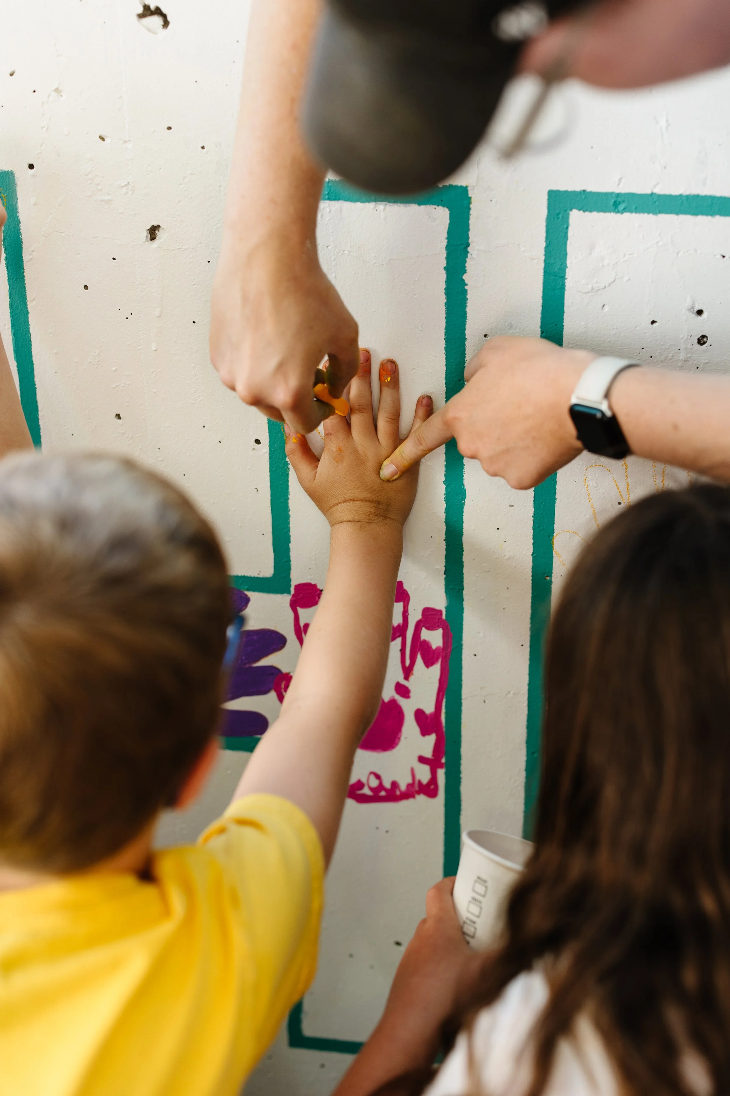 Two children and an adult painting a mural on a white wall, with one child and the adult using paintbrushes and the other child holding a cup of paint, creating colorful outlines.