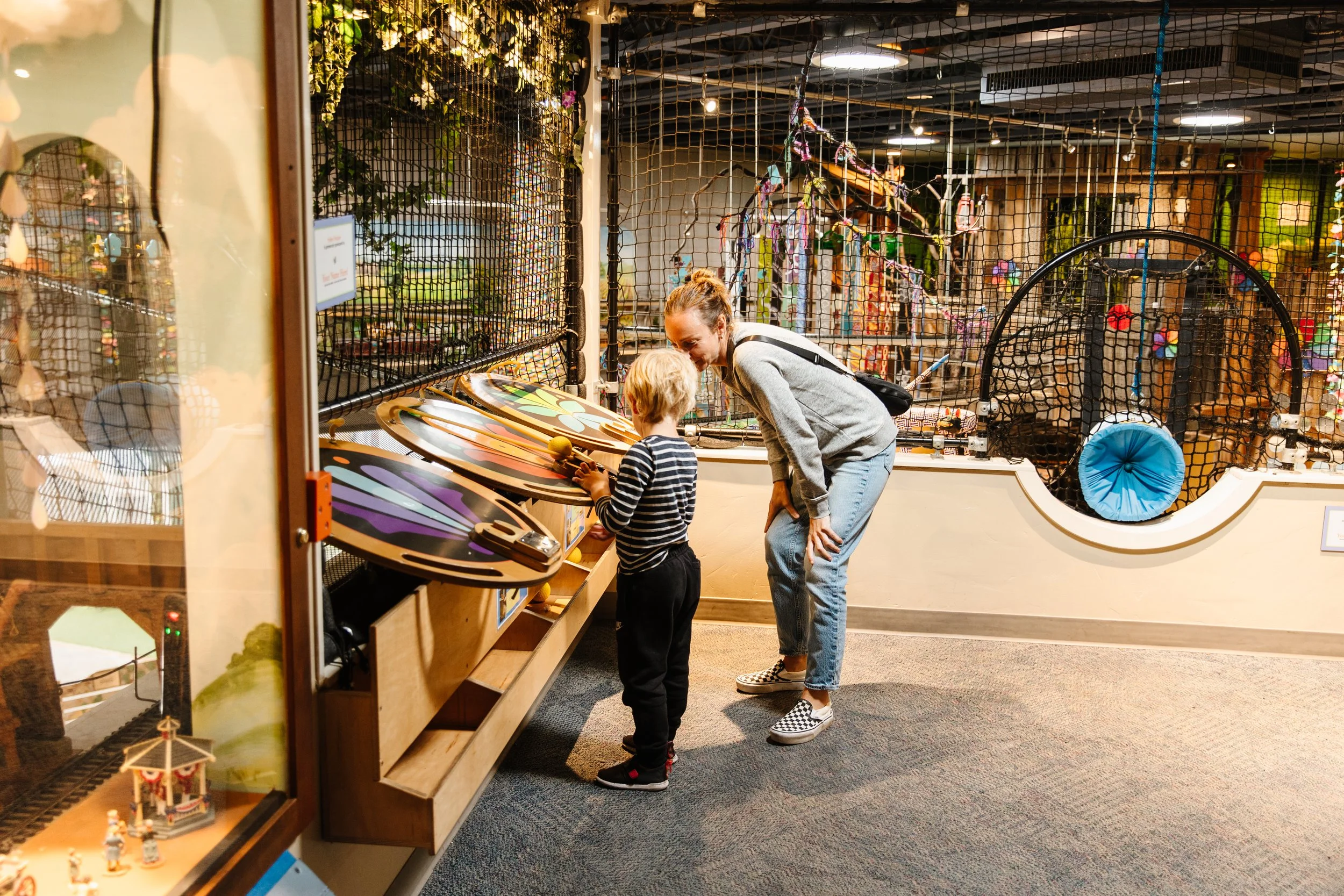 A woman and a young boy playing mini-golf at an indoor amusement facility, with colorful decorations and a small train set display visible in the background.