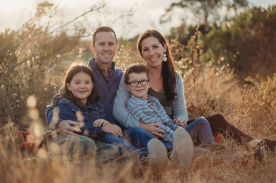 Family of four sitting together outdoors in a field with tall grass and trees in the background, smiling at the camera.