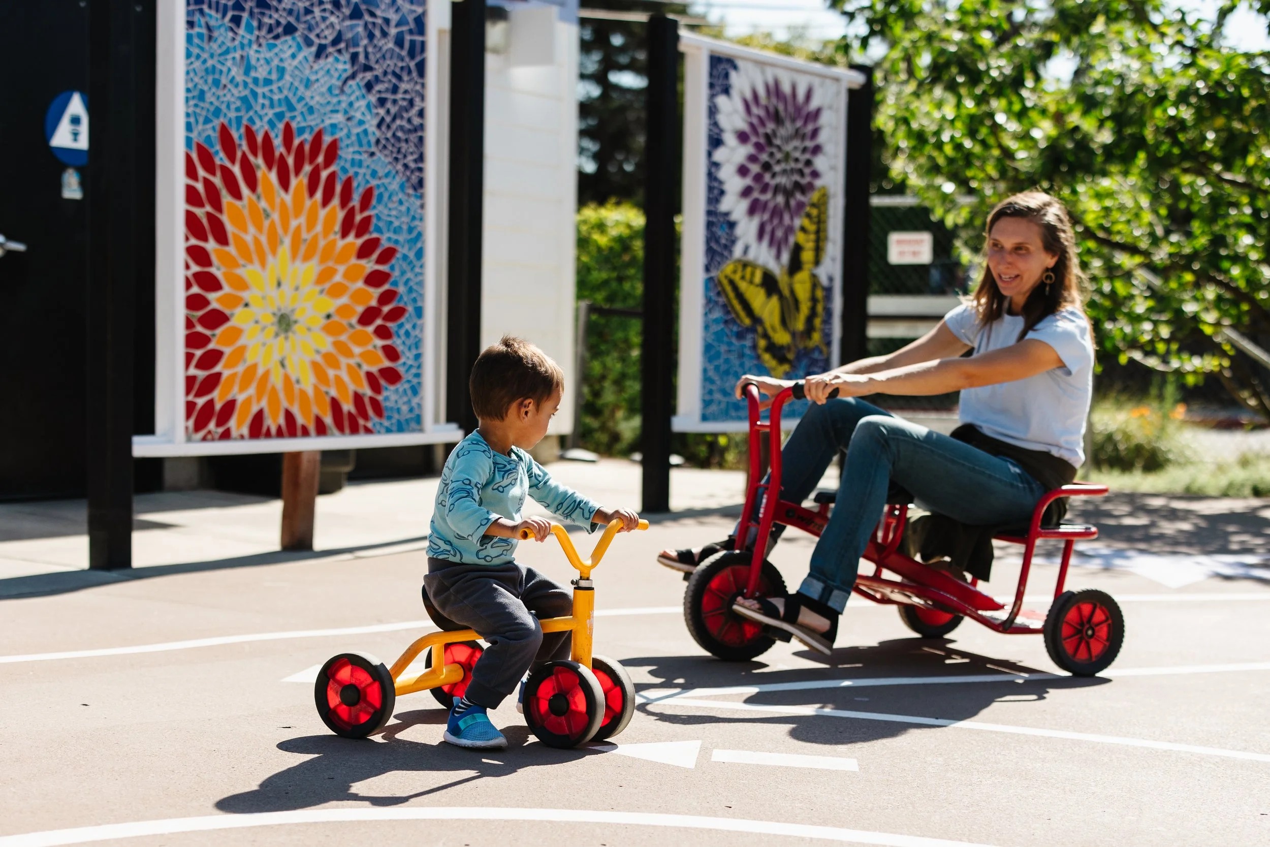 A woman and a young boy riding tricycles outdoors on a sunny day. The woman is on a red tricycle, and the boy is on a yellow tricycle. In the background, there are colorful mosaic art panels featuring a flower and a butterfly.