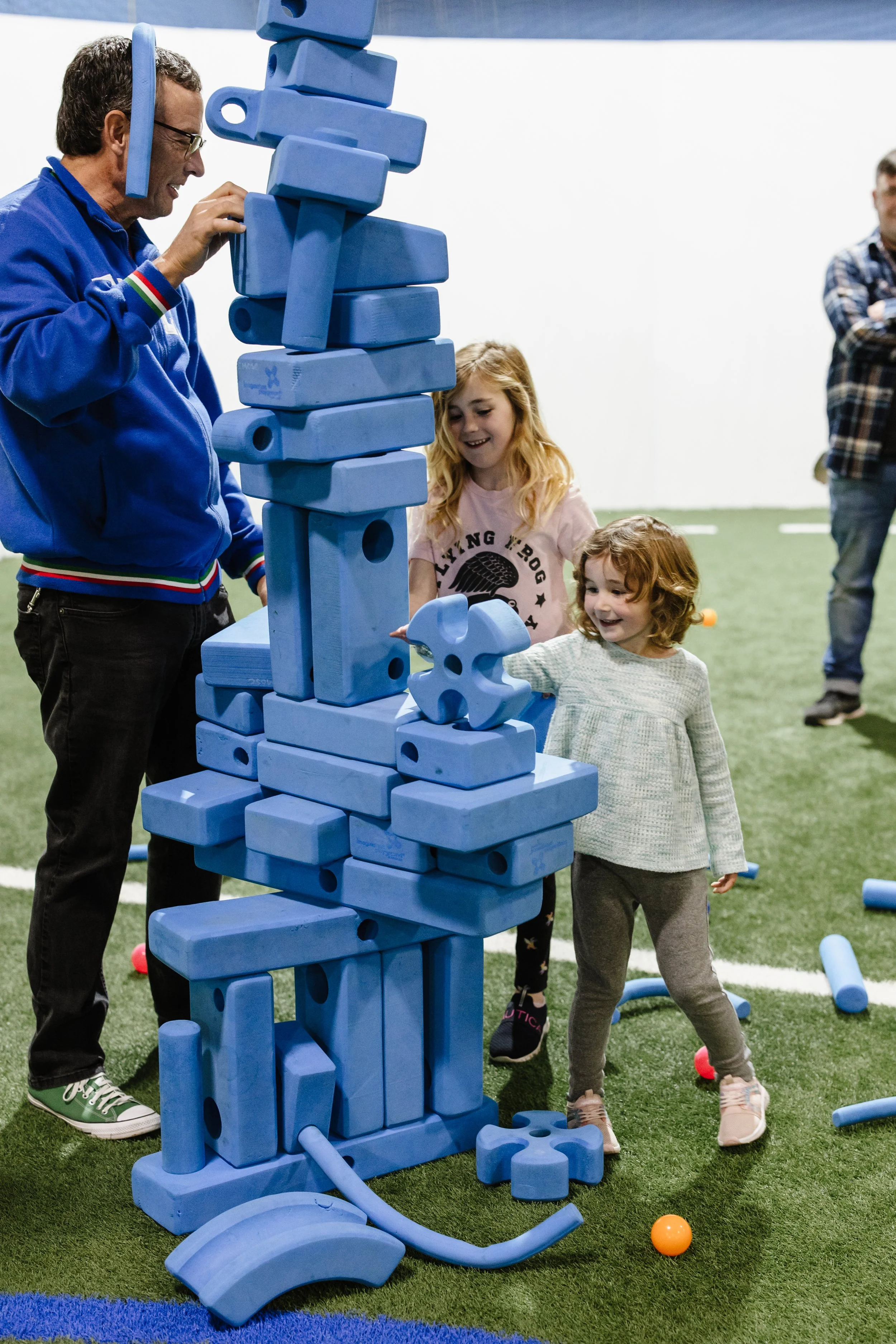 Two young girls and an adult man are playing Jenga with oversized blue blocks on an indoor field with artificial turf. The girls are smiling and looking at the tower, which is tall and slightly leaning.