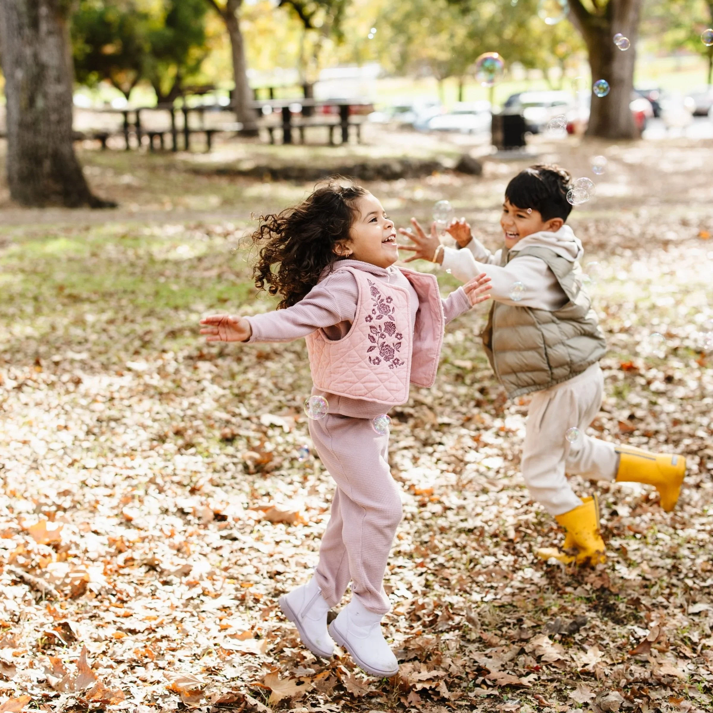 Two children playing and laughing in a park with fallen autumn leaves, surrounded by trees and sunlight, wearing warm clothes.
