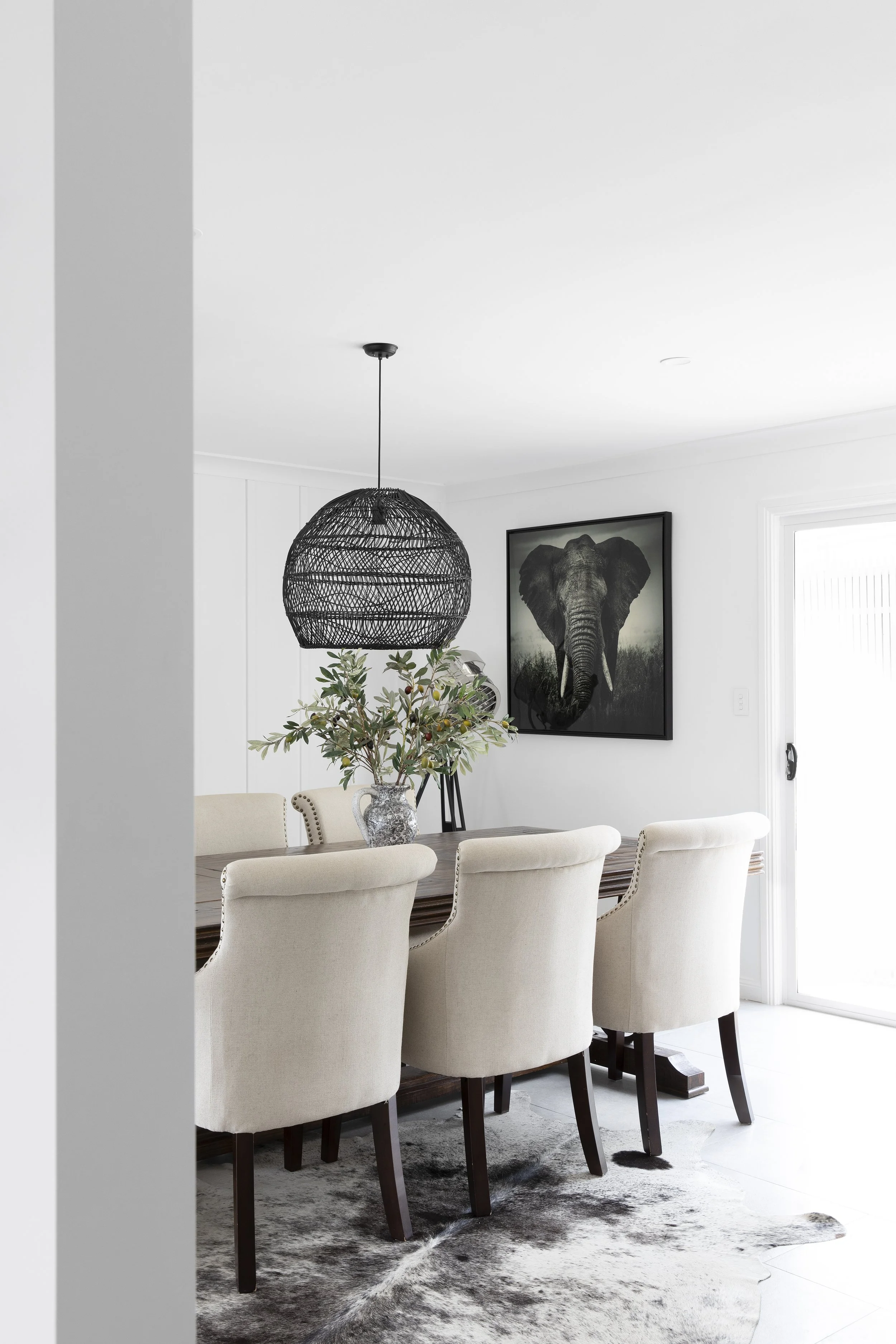 A dining room with a dark wooden table, beige upholstered chairs, a black rattan pendant light, a vase with greenery, and an elephant art photograph on a white wall near a sliding glass door.