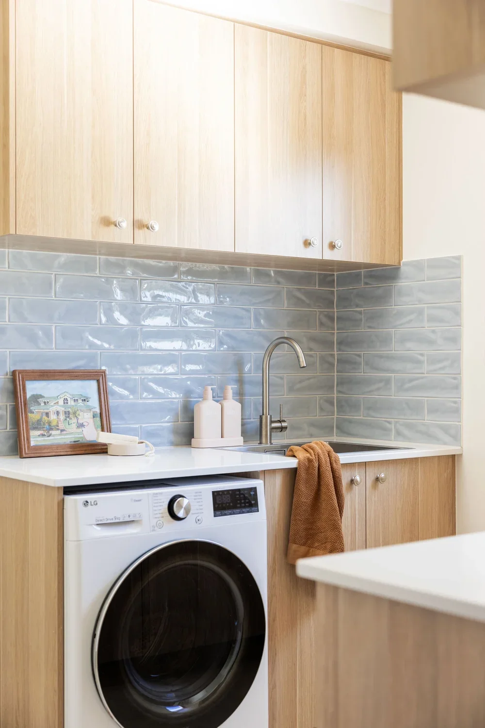 Laundry room with wooden cabinetry, gray tiled backsplash, washing machine, sink, framed picture, soap bottles, and a brown towel.