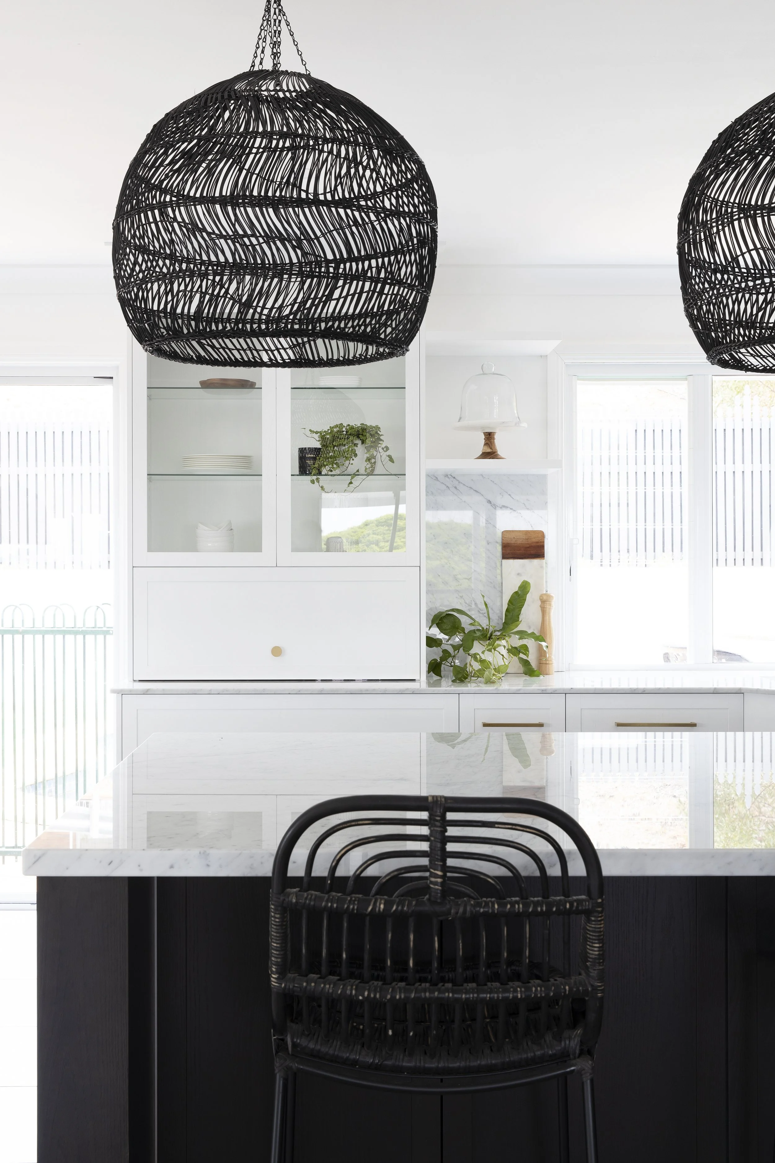 Modern kitchen with black wicker pendant lights, white cabinetry with glass doors, marble countertop, and black wicker chair at the island.