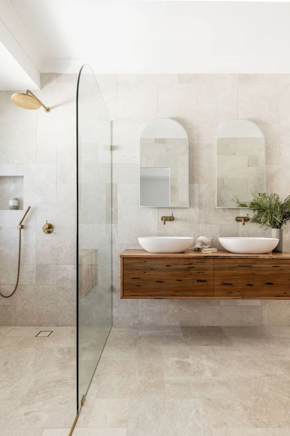 Modern bathroom with beige stone tiles, a floating wooden vanity with two vessel sinks, two large arched mirrors, a plant, and a shower area with a rainfall showerhead and a handheld shower attached to the wall.