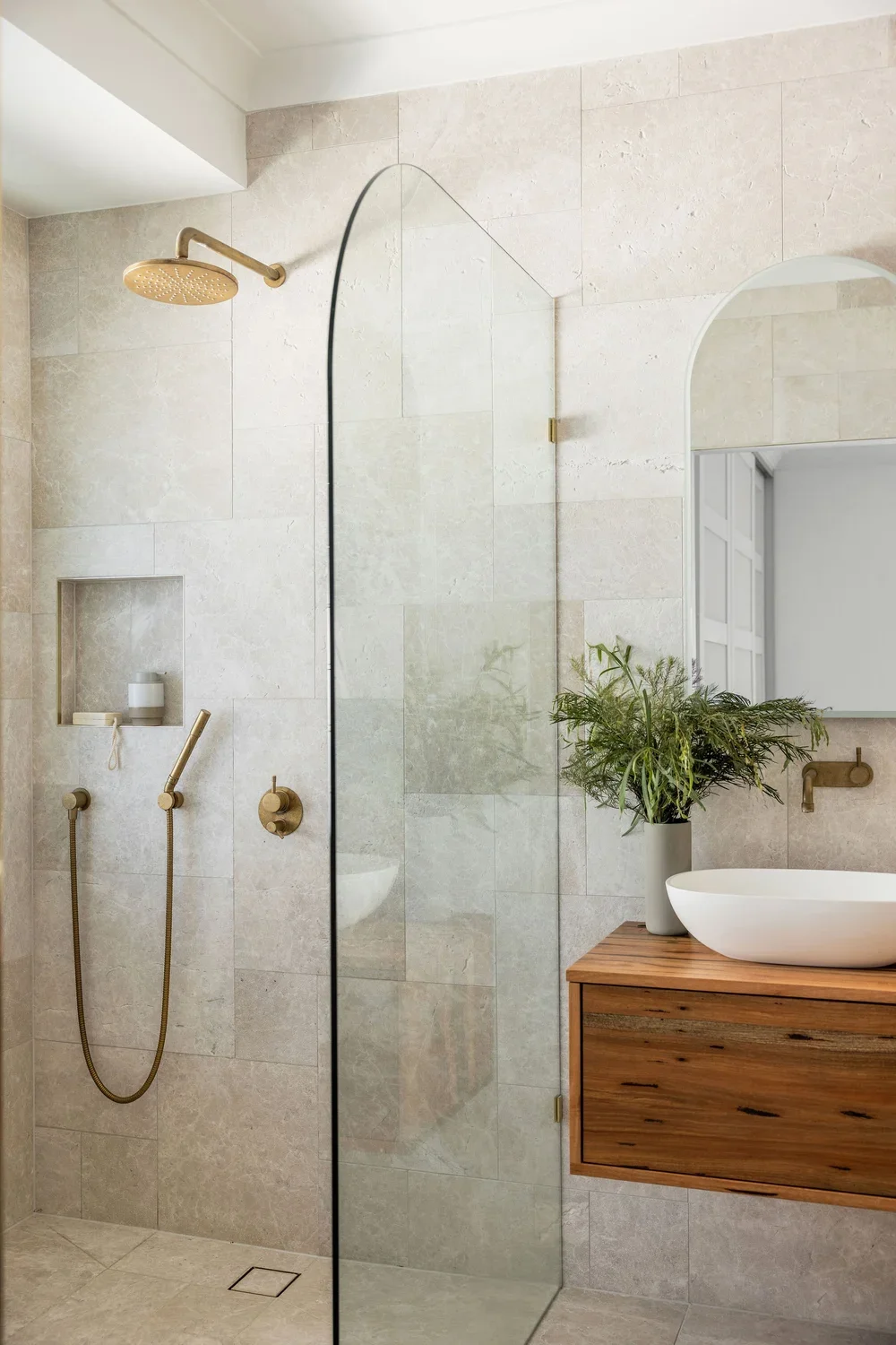 Modern bathroom with a walk-in shower, beige tiled walls, a gold showerhead, a glass partition, a wooden vanity with a white vessel sink, a large mirror, and a green plant in a gray pot.