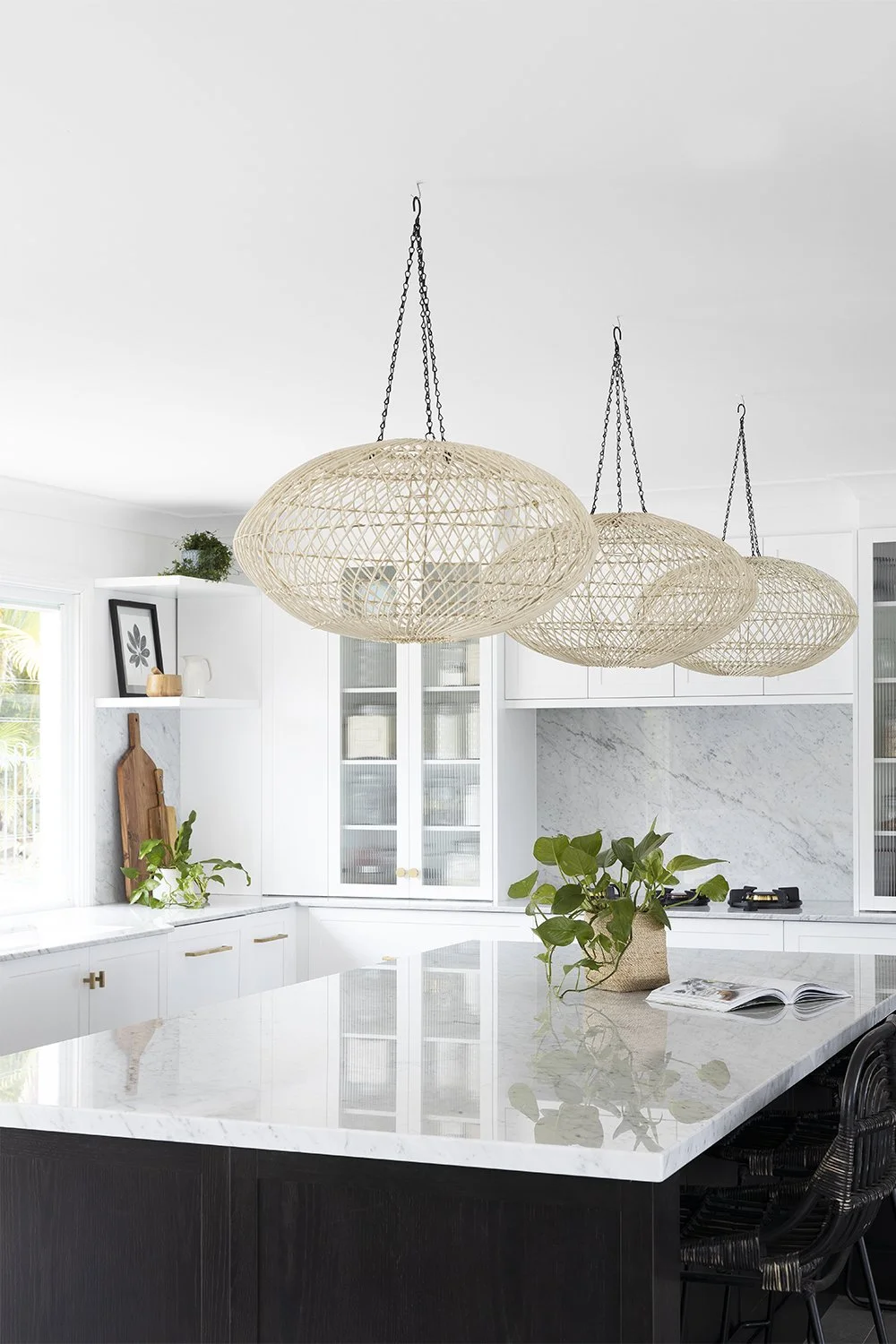 Kitchen with white cabinets, marble countertops, and three woven pendant lights hanging above a black island with a potted plant and open book.