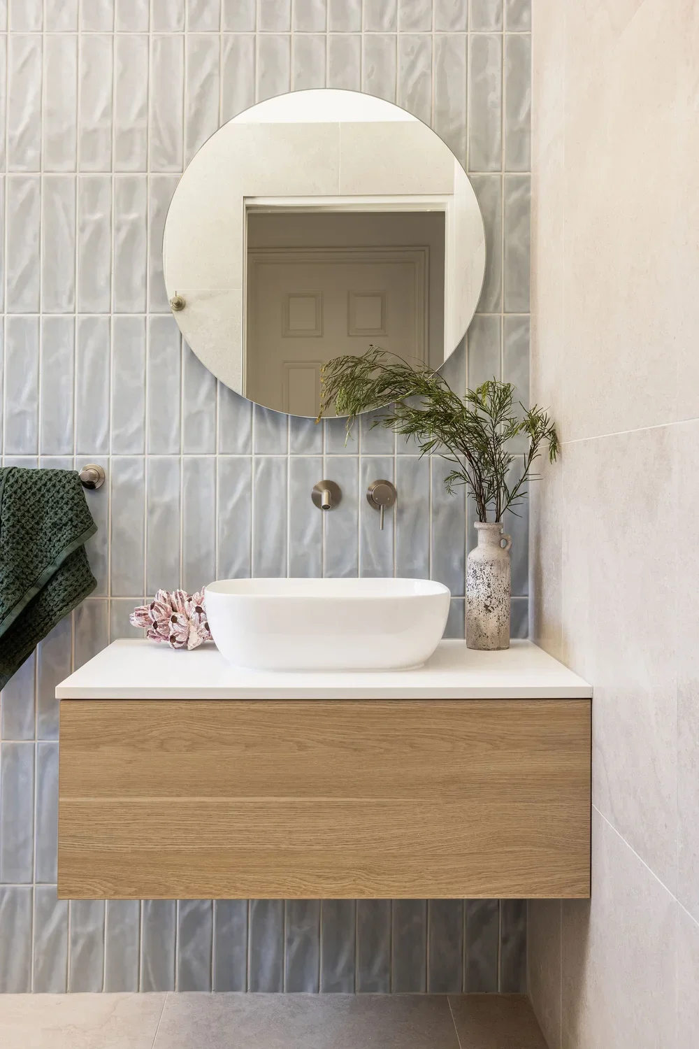 Modern bathroom with a round mirror, a white vessel sink, wooden vanity, green towel, decorative seashell, vase with greenery, and tiled wall.