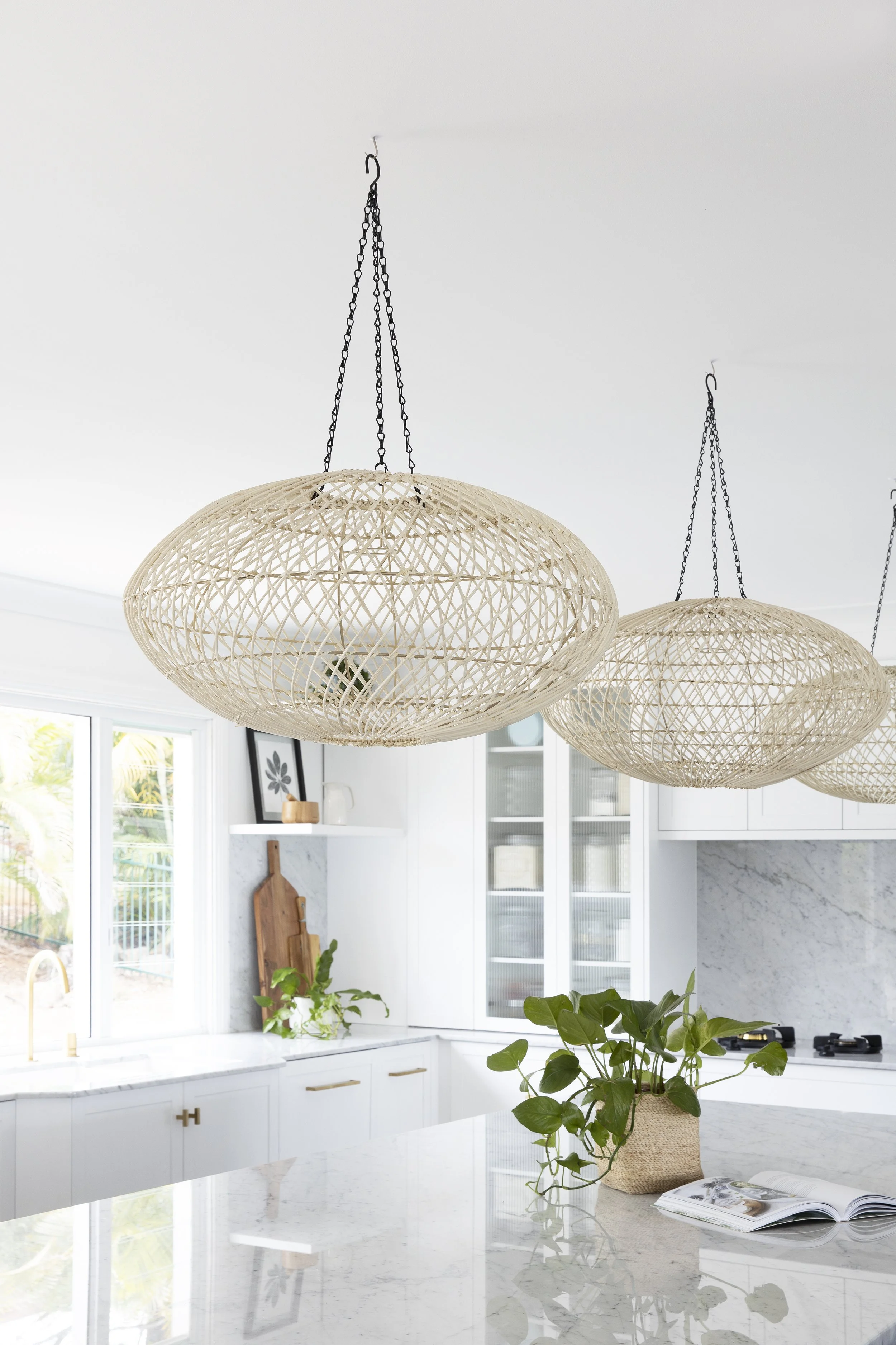 Bright modern kitchen with white cabinetry, marble countertops, and a marble backsplash. Decor includes framed art, a cutting board, a potted plant, and an open magazine. Three woven pendant lights hang from the ceiling.