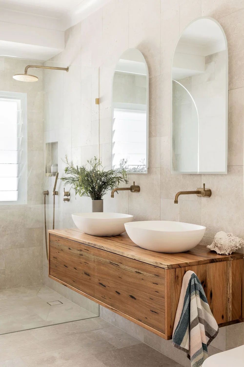 Modern bathroom with double vessel sinks on a wooden vanity, two oval mirrors, brass faucets, a plant, and a shower area with a rainfall showerhead.