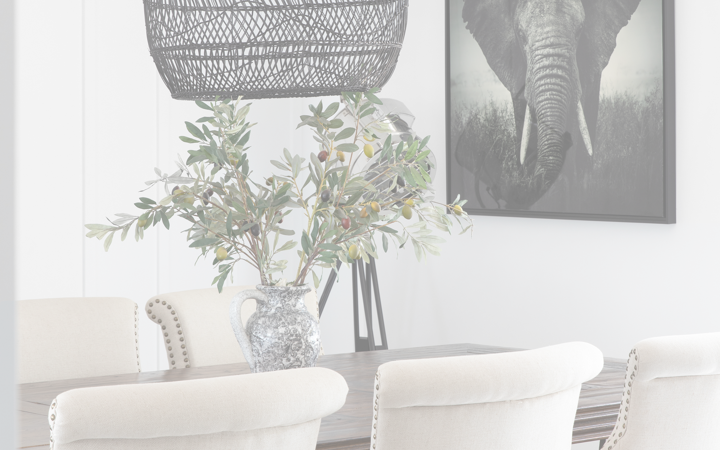 A dining room with a table, four upholstered chairs, a large vase with greenery and olives, a large black wire hanging lamp, and a framed elephant photograph on the wall.