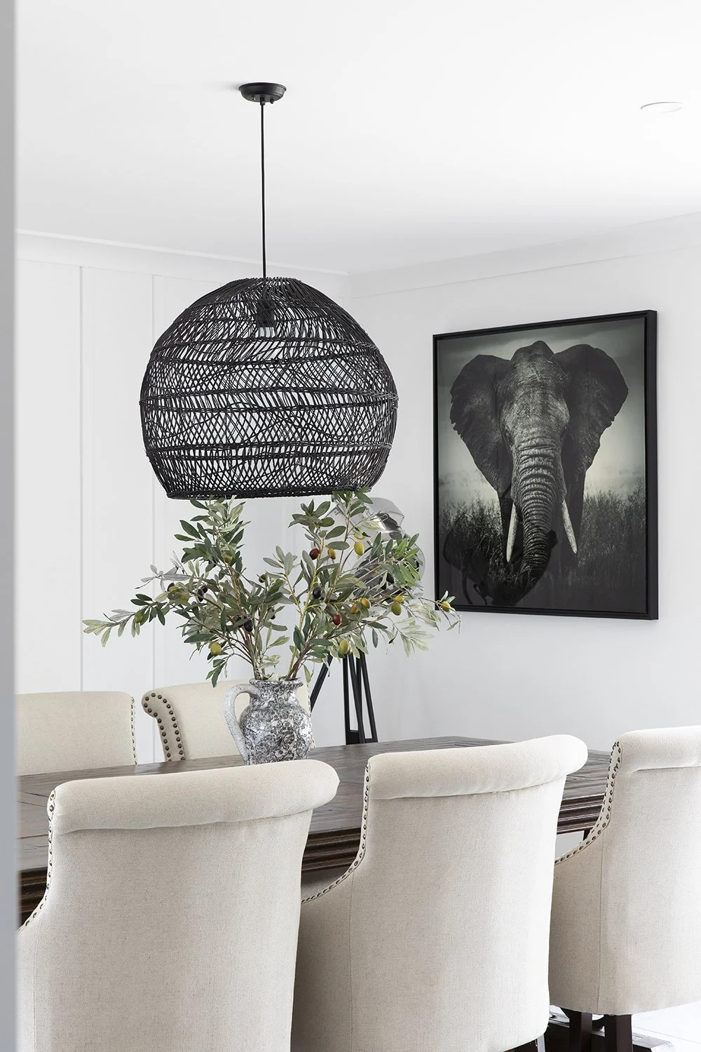 A dining room with a black wicker pendant light, a wooden table, beige upholstered chairs, a decorative vase with green foliage, and a framed black-and-white elephant photograph on the wall.