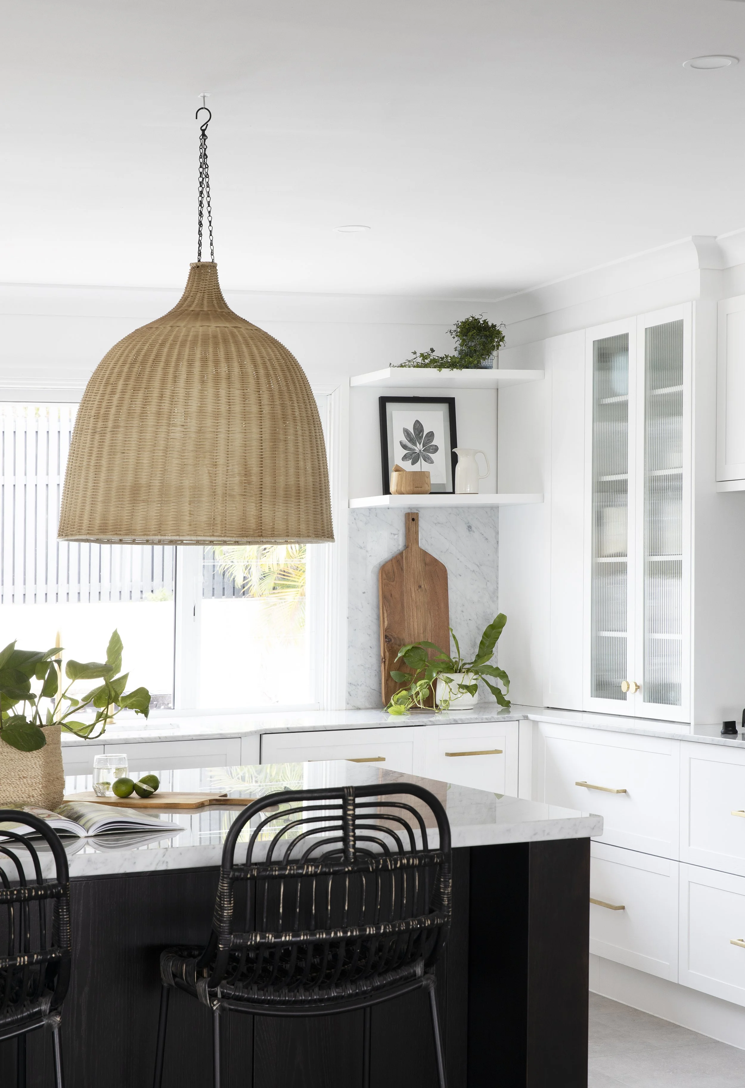 Modern kitchen with white cabinets, marble countertops, black chairs, green plants, wicker pendant light, and open shelving with decor.
