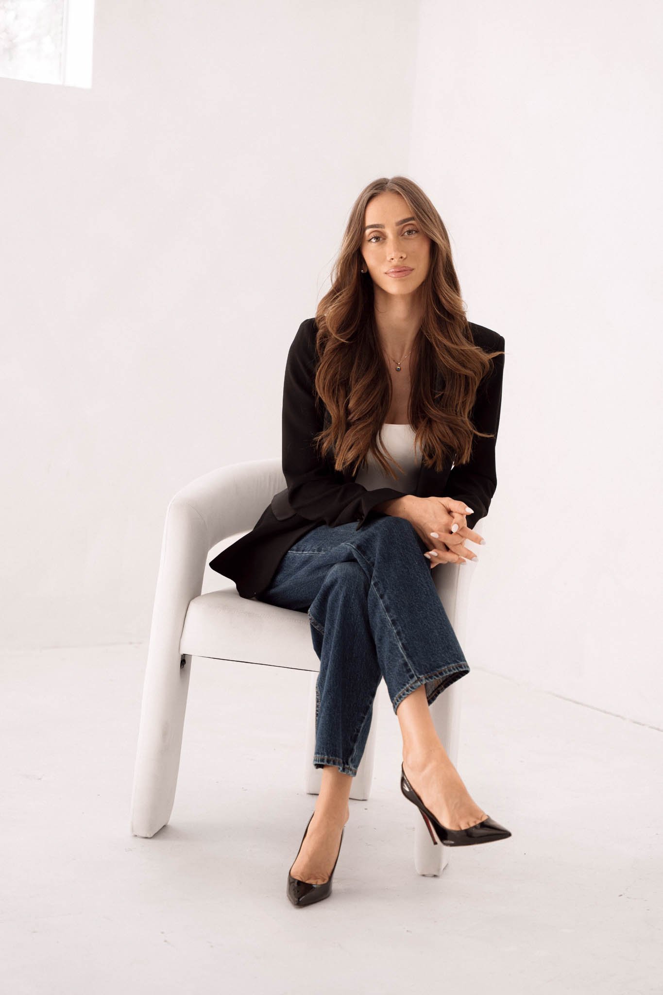 A woman with long brown hair sits on a white chair against a white background. She wears a black blazer, white top, blue jeans, and black high heels.