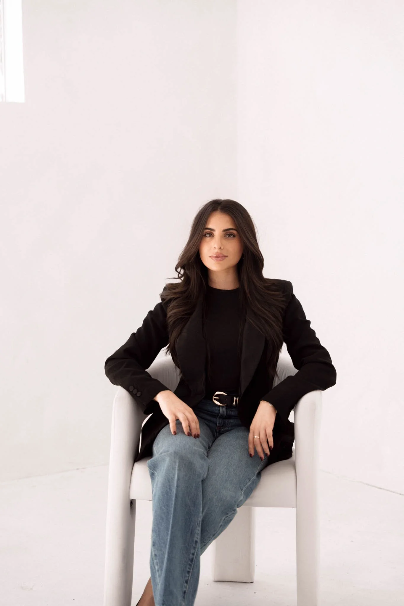 A woman with long, dark hair, dressed in a black blazer and jeans, is sitting on a white chair in a minimalist white room.