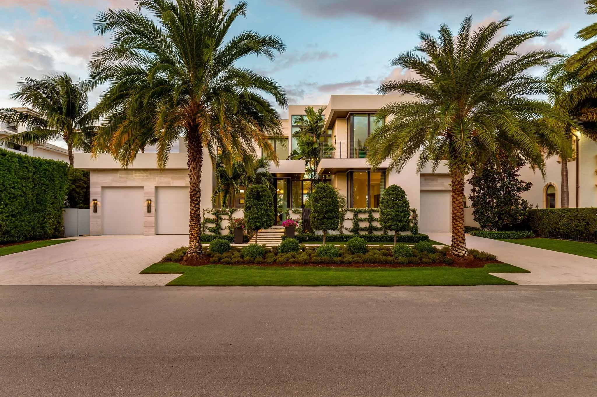 front view of a modern house with white walls, large glass windows, and a flat roof, surrounded by tall palm trees and neatly trimmed bushes, with a driveway leading to garages, under a partly cloudy sky.