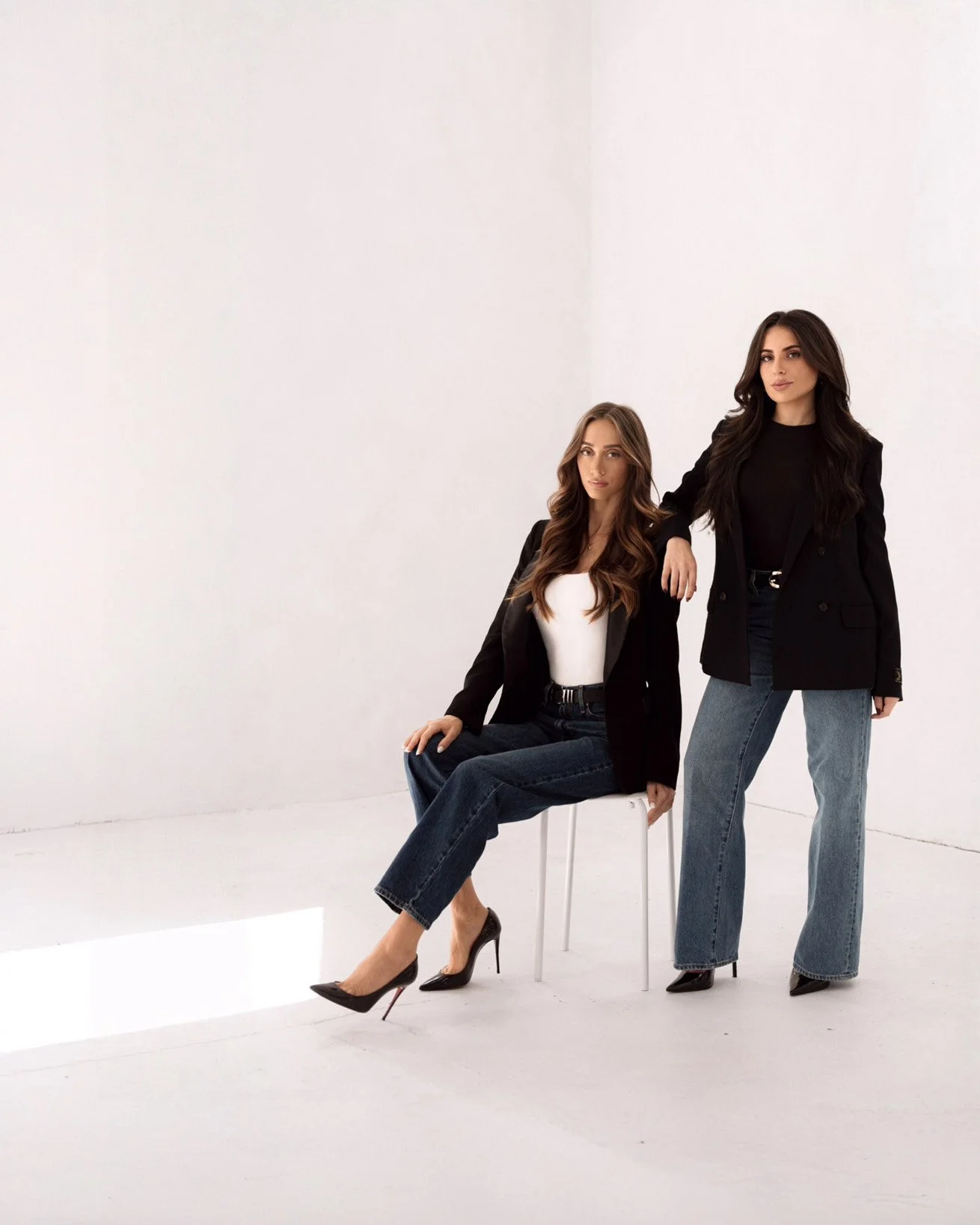Two women in black blazers and jeans posing in a white studio, one seated on a white chair and the other standing with her hand on the seated woman's shoulder.