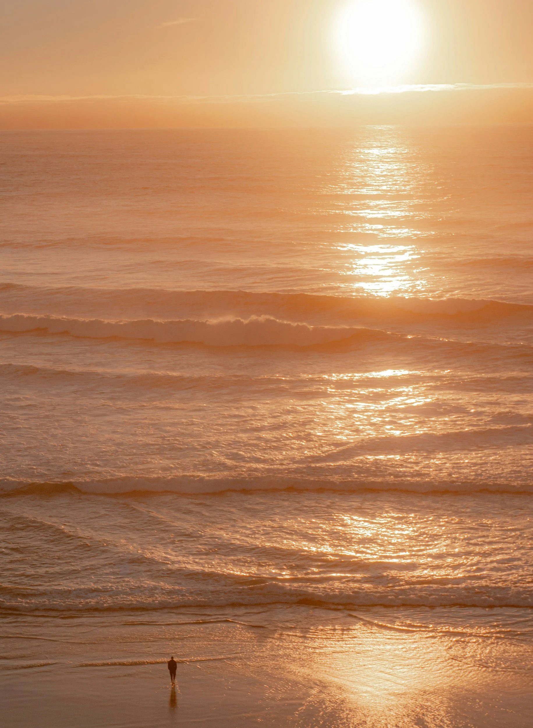 A person standing on the beach during sunset, with the ocean reflecting the orange and gold colors of the sky.