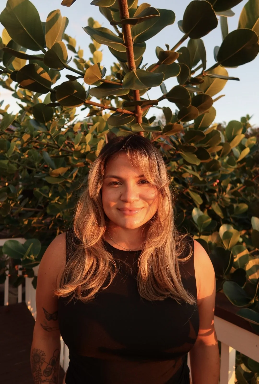 A woman with blonde hair smiling outdoors during sunset, standing in front of green leafy plants on a patio with a white railing.