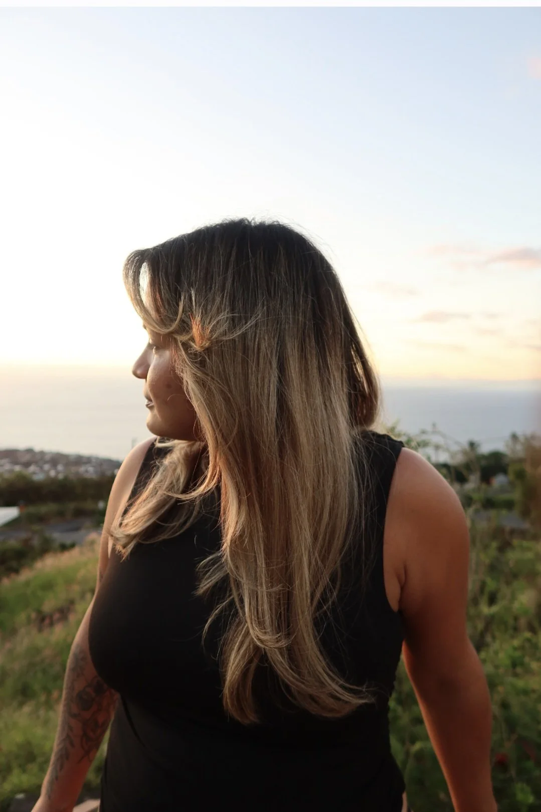 A woman with long wavy hair wearing a black sleeveless top, standing outdoors during sunset with a scenic view of the ocean and sky in the background.