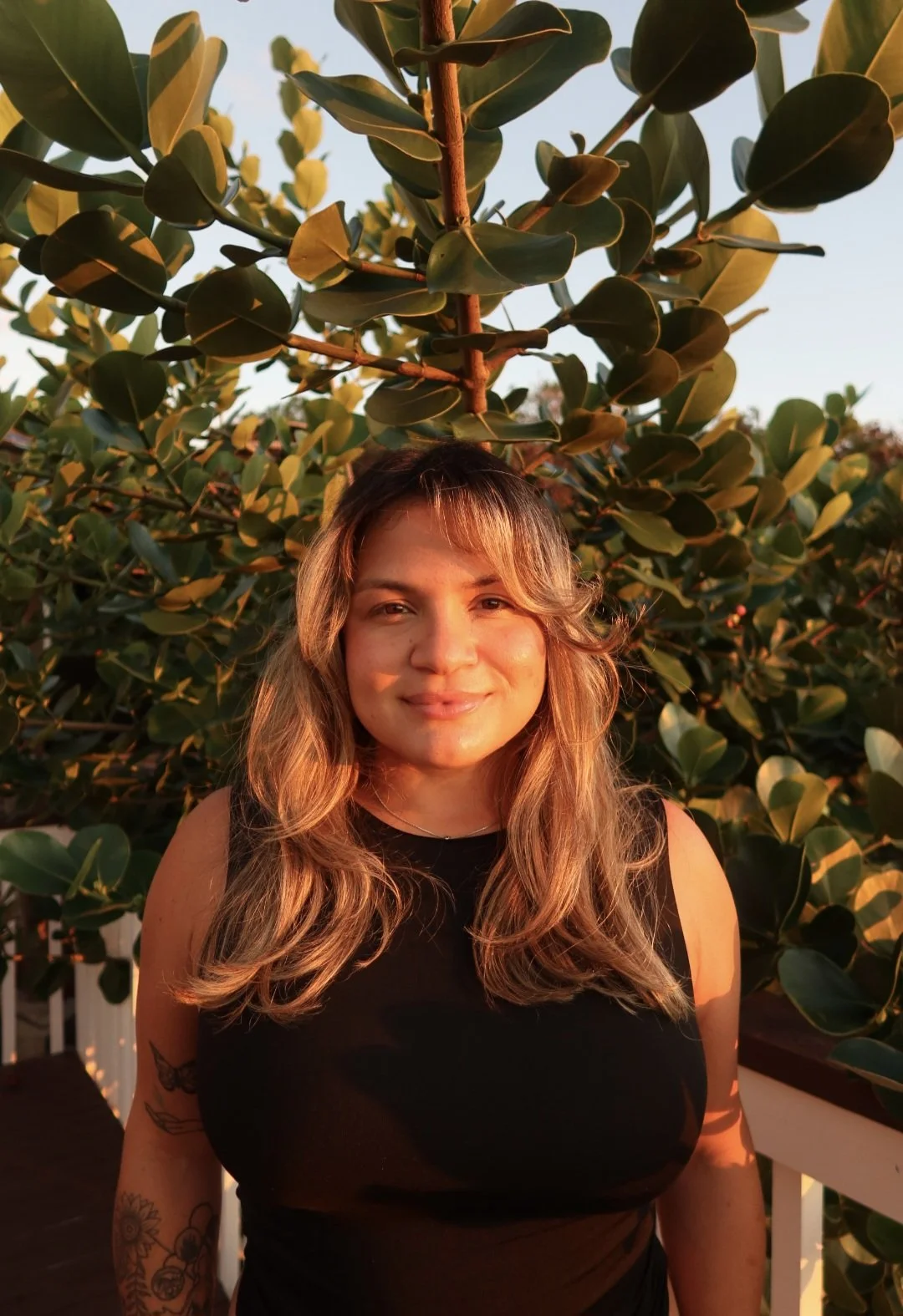 A woman with long wavy hair, wearing a black sleeveless top, smiling in sunset light against a backdrop of green leaves from a large plant or tree.
