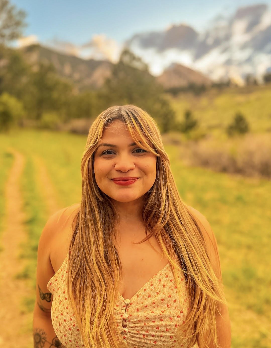 A smiling woman with long blonde hair in a field of green grass with hills and mountains in the background.
