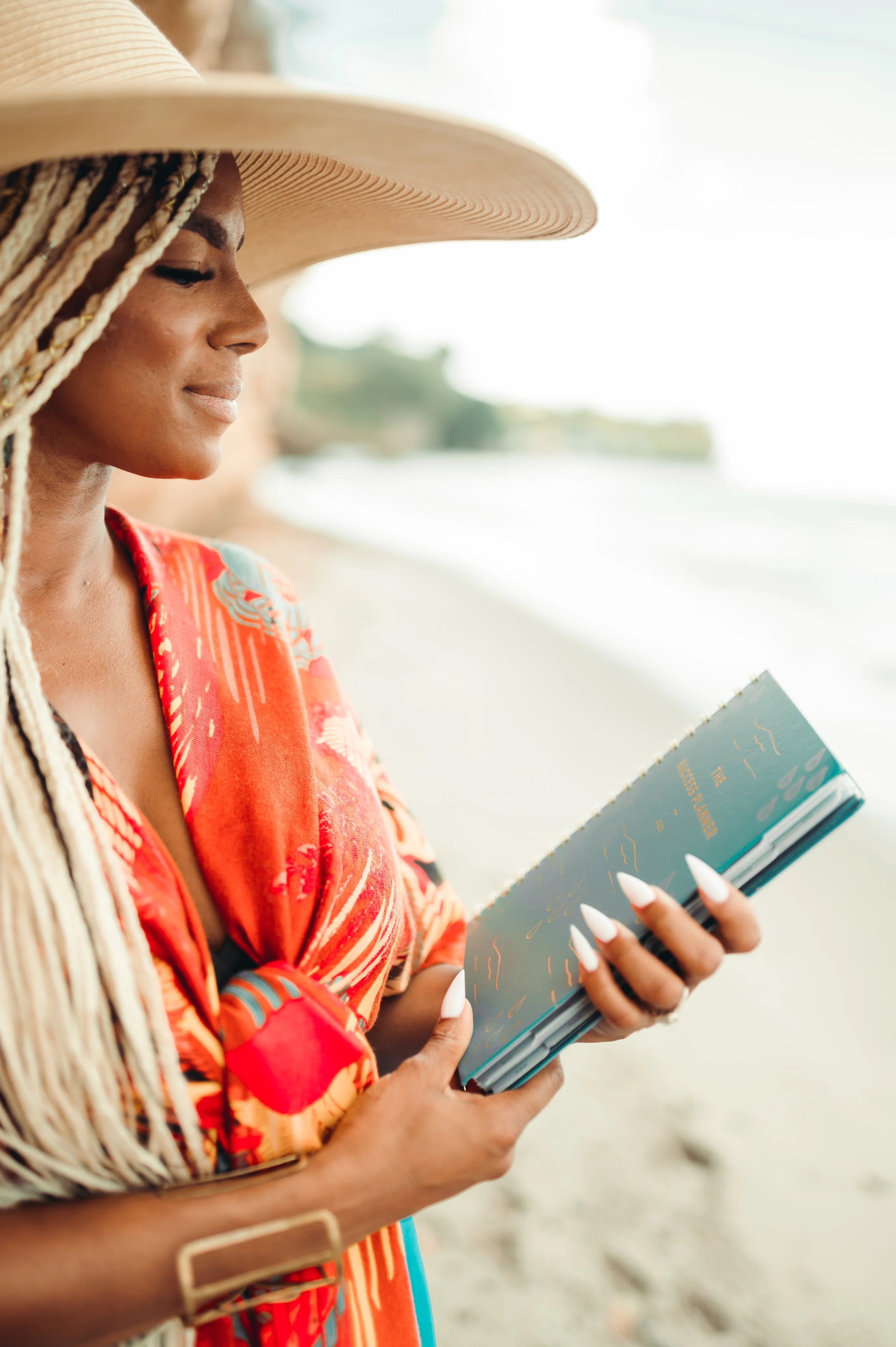 Woman with long braided hair wearing a wide-brimmed hat and colorful dress reading a book on a beach.